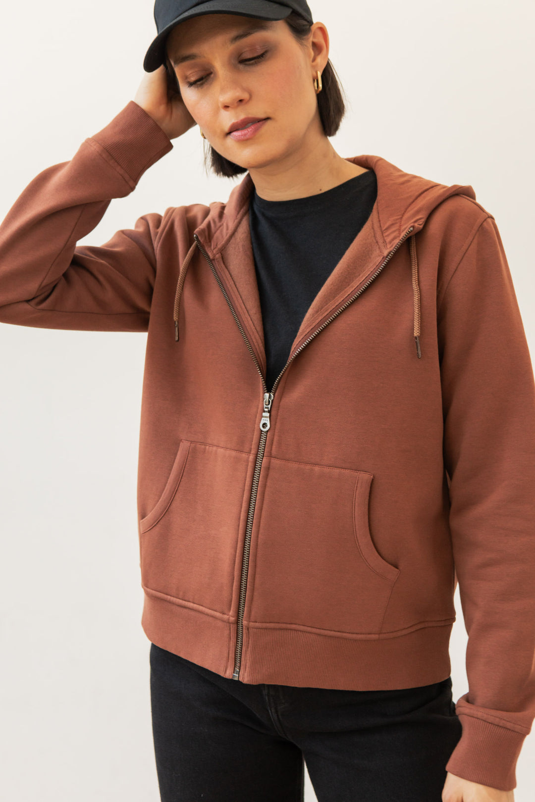 A person in the Bridge & Burn Mason Hoodie in Burnt Umber, layered over a black shirt, pants, and cap, stands with one hand on their head and eyes closed against a plain light background.