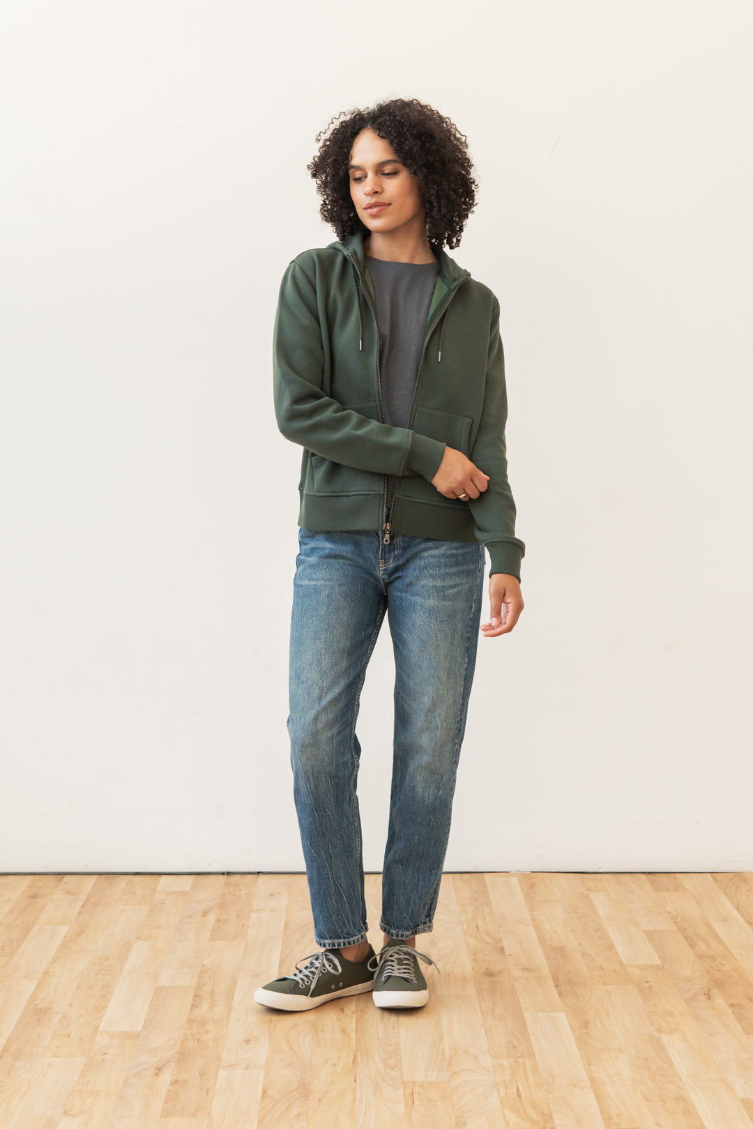 A person with curly hair wears the Bridge & Burn Mason Hoodie in Forest Night, a gray t-shirt, blue jeans, and green sneakers while standing on a light wood floor against a plain white wall.