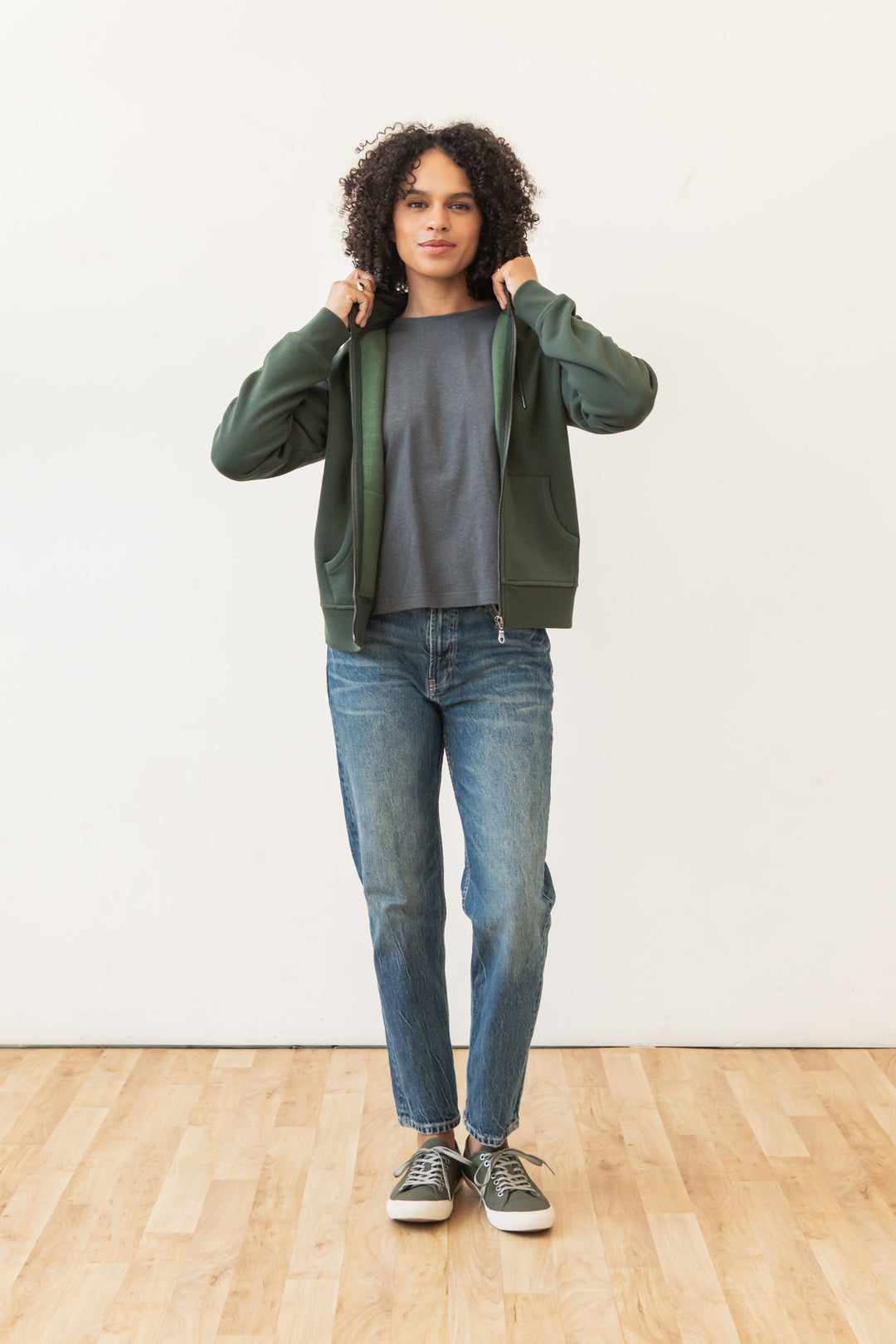 A person with curly hair stands on a wooden floor against a plain white wall, wearing the Bridge & Burn Mason Hoodie in Forest Night over a gray t-shirt, blue jeans, and gray sneakers, holding the hoodie's collar with both hands.