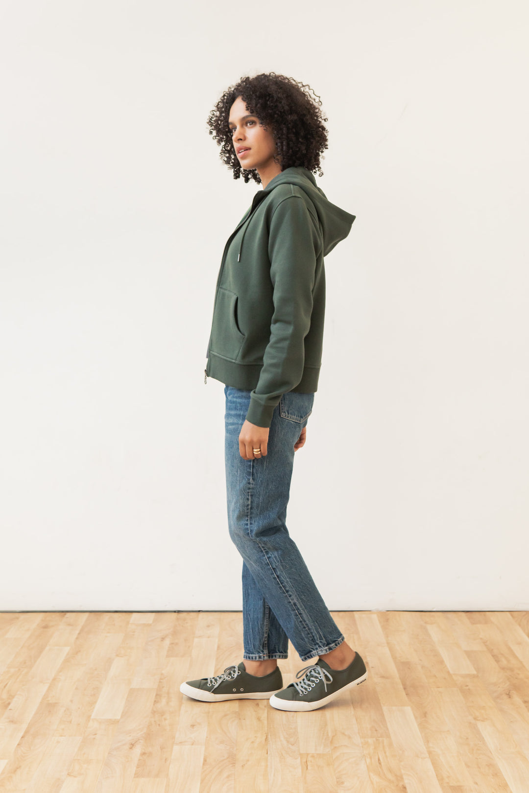 A person with curly hair wears the Bridge & Burn Mason Hoodie in Forest Night, paired with blue jeans and dark sneakers, standing sideways on a light wooden floor against a plain white wall.