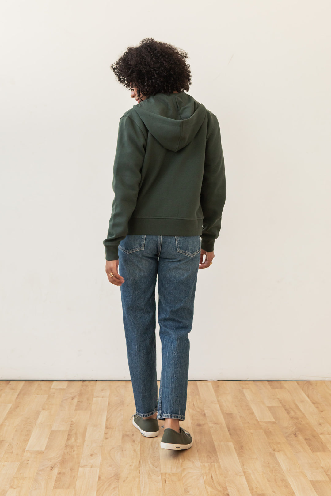 A person with curly hair stands facing a white wall on a light wood floor, wearing the Bridge & Burn Mason Hoodie in Forest Night, blue jeans, and gray slip-on shoes.
