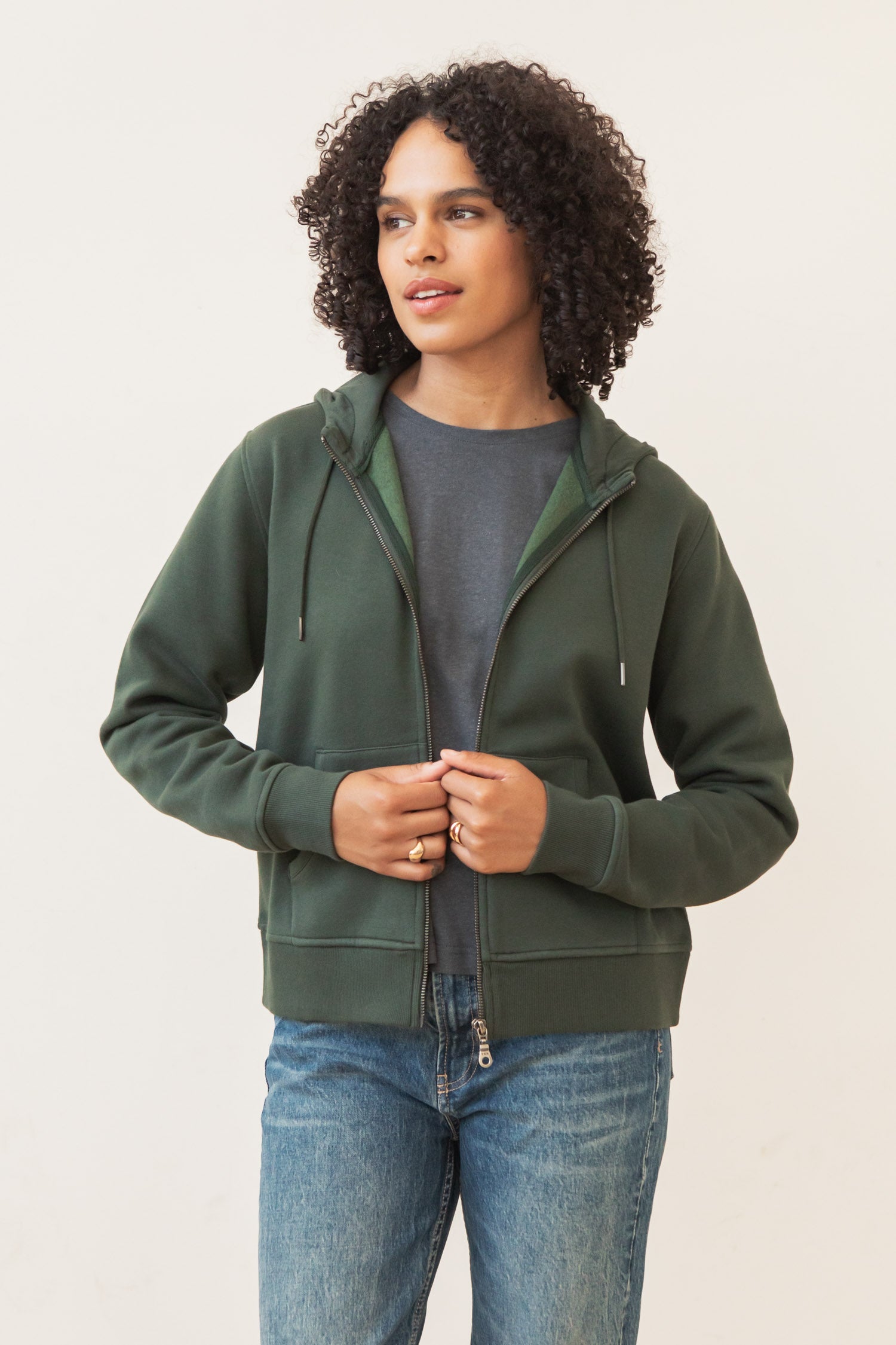 A person with curly hair wears the Bridge & Burn Mason Hoodie in Forest Night over a gray t-shirt and blue jeans, standing against a plain light background and looking to the side while holding the hoodie zipper.