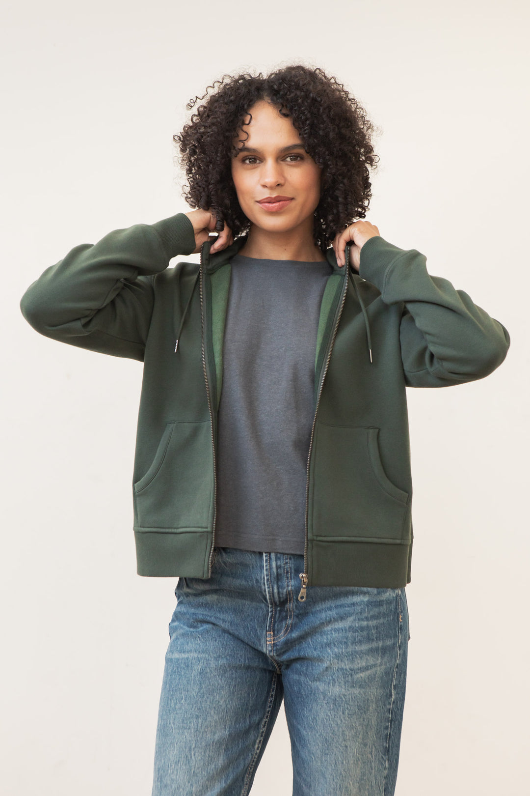 A person with curly hair wears the Bridge & Burn Mason Hoodie in Forest Night over a gray shirt and blue jeans, holding the hoodie by the collar and facing the camera against a plain light background.