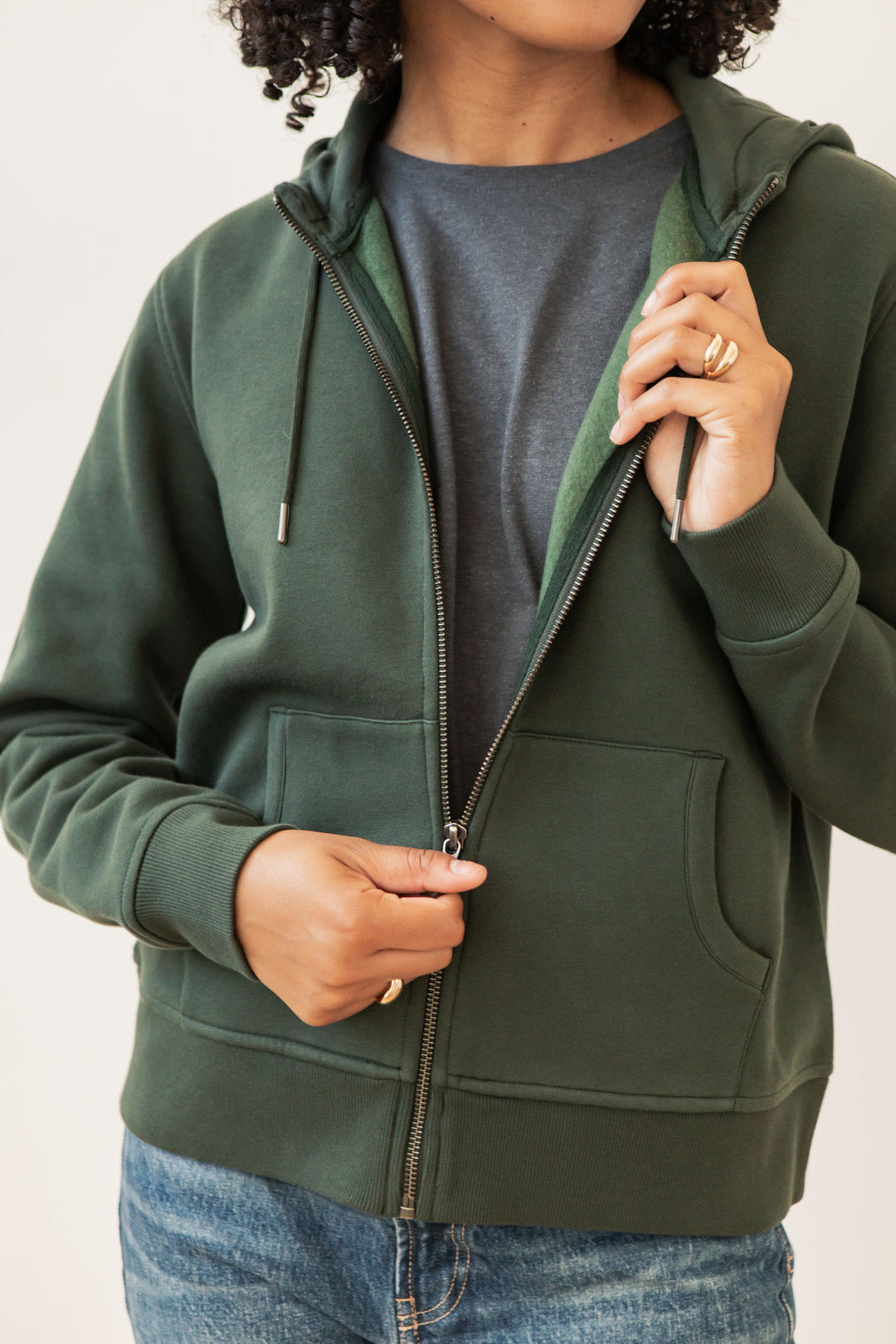 A person with chin-length curly hair and gold rings unzips the Bridge & Burn Mason Hoodie in Forest Night, layered over a gray shirt and paired with denim jeans. The background is plain and light-colored.