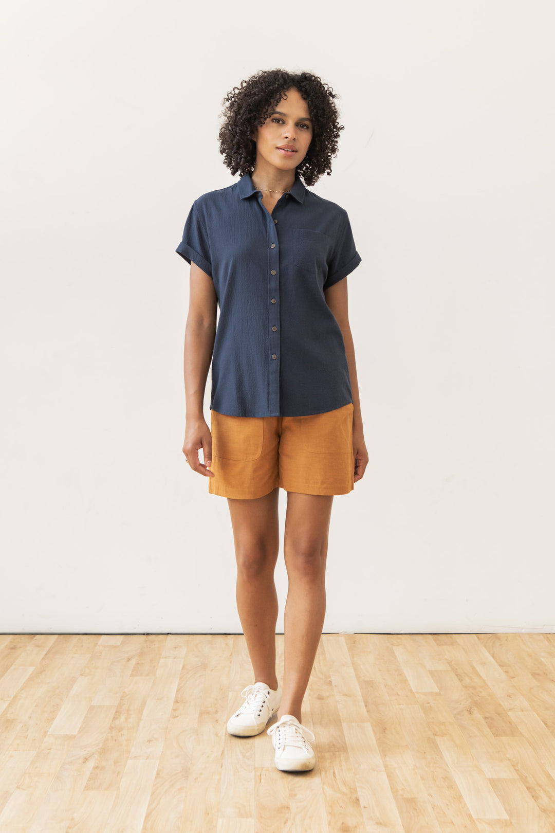 A woman with curly hair stands indoors on a wooden floor, wearing the Bridge & Burn Bea Shirt in Navy Seersucker, mustard-colored shorts, and white sneakers against a plain white wall.