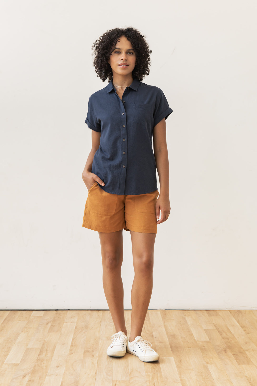 Indoors on a wooden floor, a person with curly hair and one hand in their pocket wears the Bridge & Burn Bea Shirt in Navy Seersucker, paired with mustard shorts and white sneakers, looking at the camera.