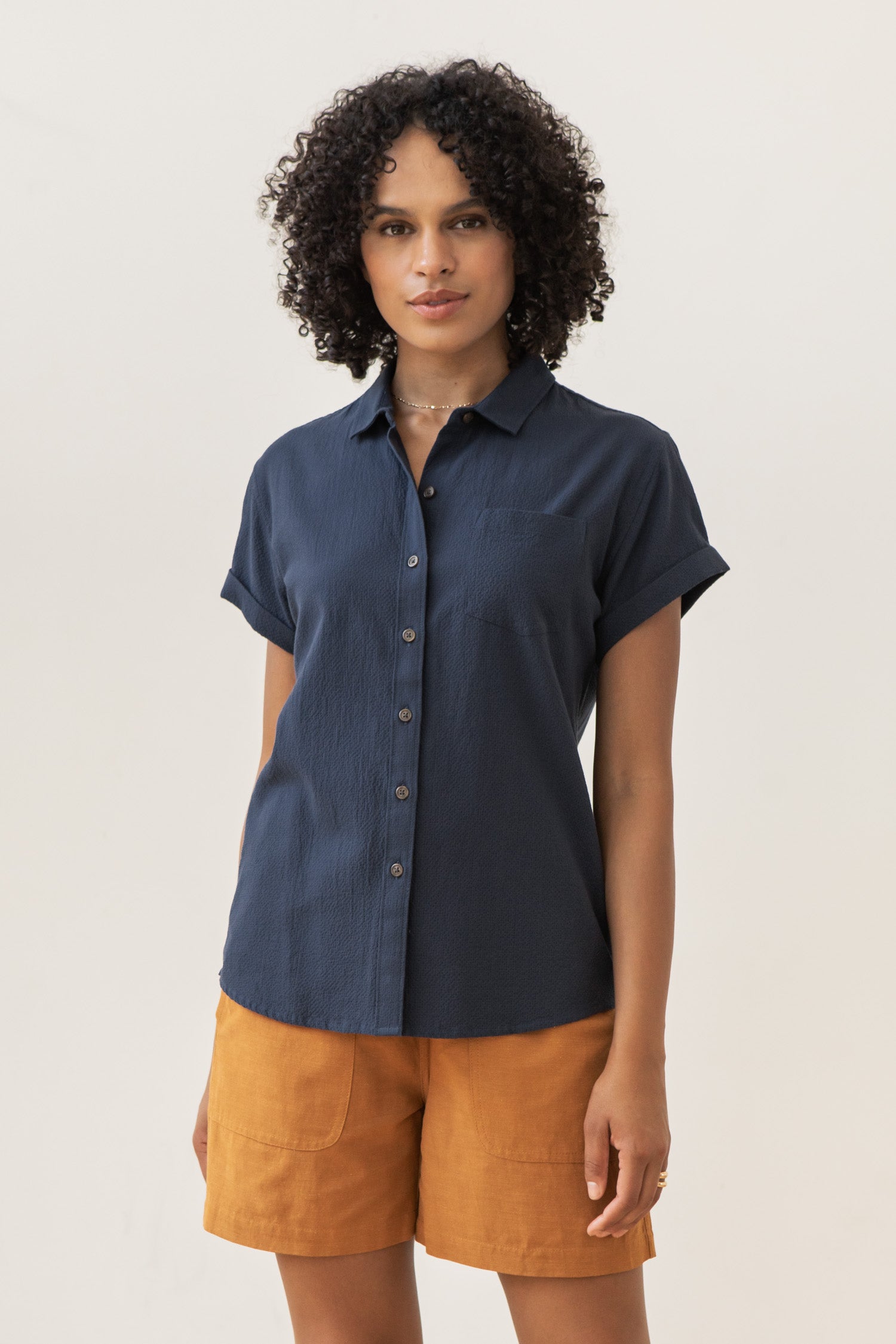 A person with curly hair wears the Bridge & Burn Bea Shirt in Navy Seersucker and mustard yellow shorts, standing against a plain light background and looking at the camera.