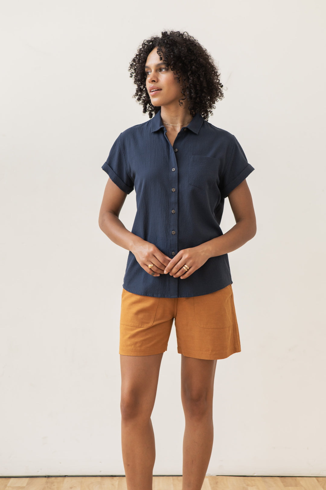 A person with short curly hair and a neutral expression stands indoors against a plain wall, wearing the Bridge & Burn Bea Shirt in navy seersucker and mustard-colored shorts.