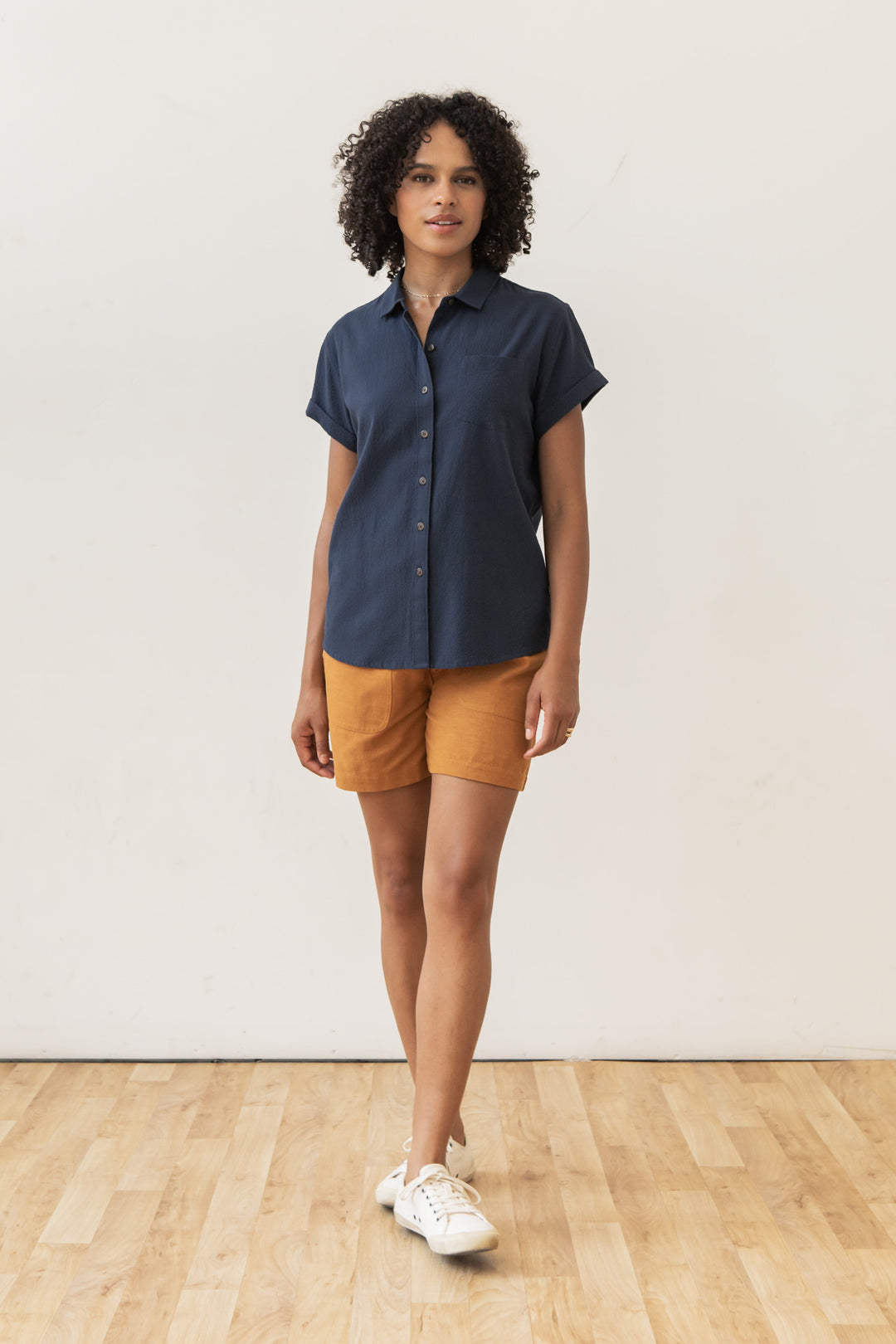 A person stands indoors on a wood floor, wearing the Bridge & Burn Bea Shirt in Navy Seersucker, orange-brown shorts, and white sneakers against a plain white wall.