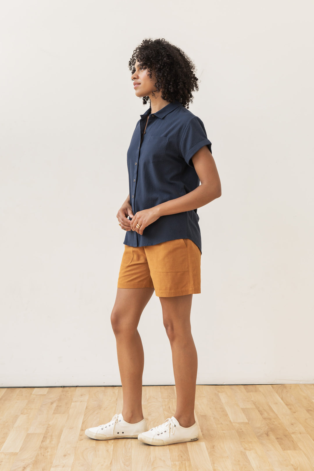 A person with curly hair stands in profile on a wooden floor against a white wall, wearing the Bridge & Burn Bea Shirt in Navy Seersucker, rust-colored shorts, and white sneakers.