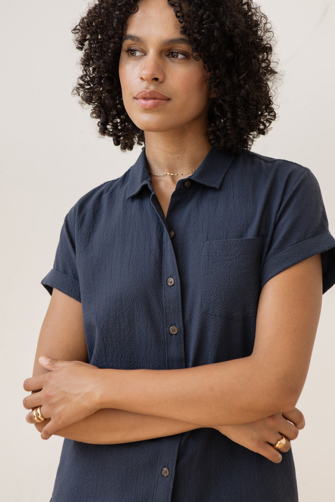 A woman with curly hair, wearing the Bridge & Burn Bea Shirt in Navy Seersucker, stands with her arms crossed and gazes to the side with a neutral expression.