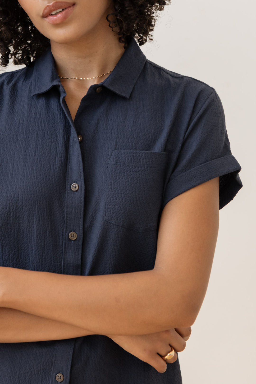 A woman with curly hair wears the Bridge & Burn Bea Shirt in Navy Seersucker, styled with a delicate necklace and ring. She stands with arms crossed, facing partially away from the camera.