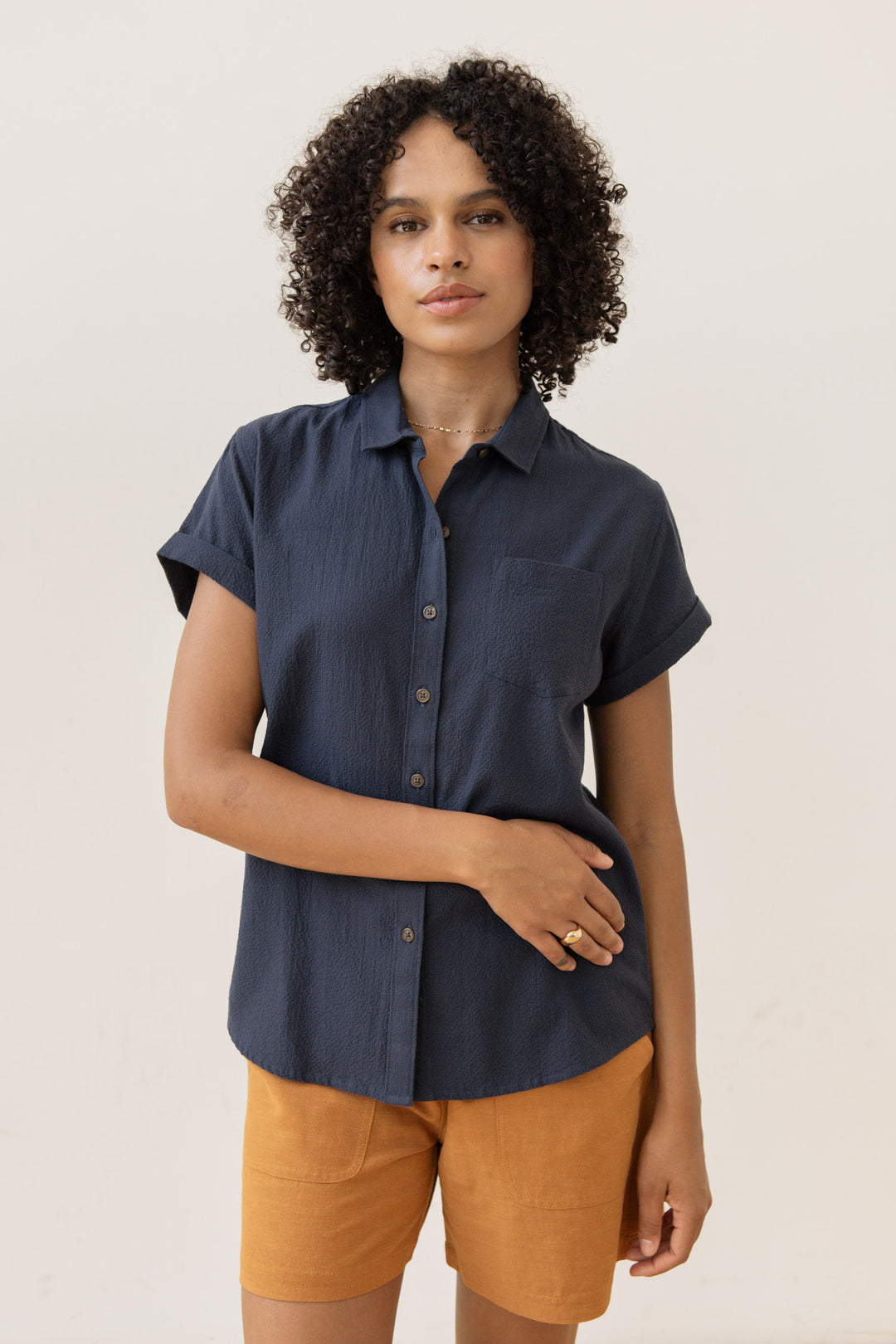 A person with curly hair wears the Bridge & Burn Bea Shirt in Navy Seersucker and mustard shorts, standing against a plain light background and calmly gazing at the camera with one hand resting on their stomach.