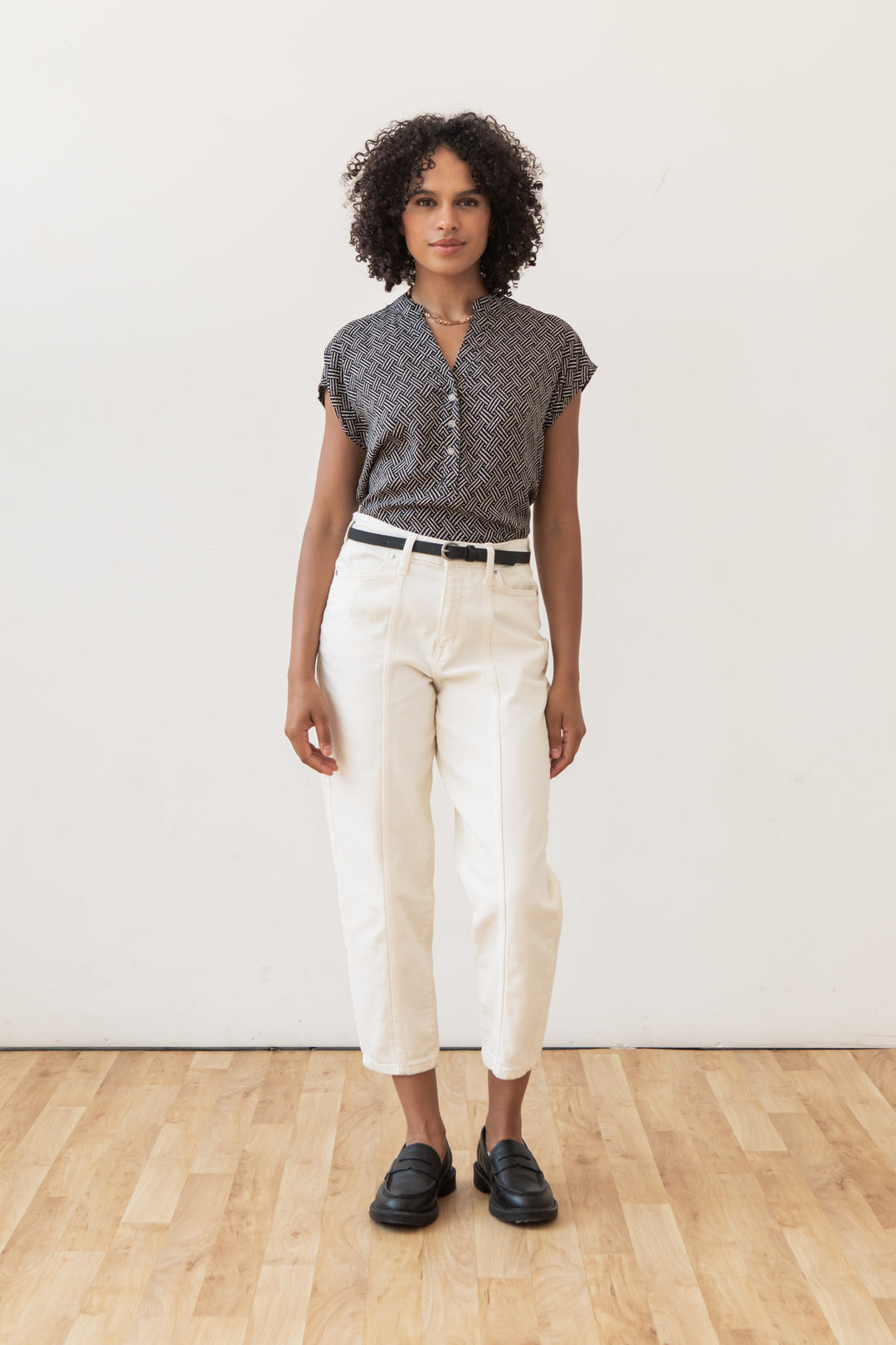 A woman with curly hair stands indoors on a wooden floor, wearing the Bridge & Burn Ladd Shirt in Black Geo, white high-waisted pants, a black belt, and black loafers against a plain white wall.