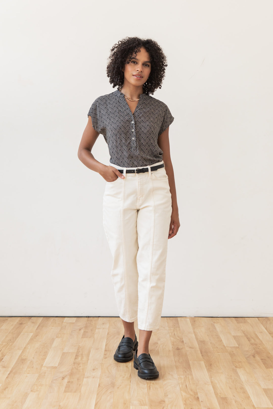 A woman with curly hair stands on a wooden floor, wearing the Bridge & Burn Ladd Shirt in Black Geo, white high-waisted pants with a black belt, and black loafers. One hand is in her pocket as she looks forward.