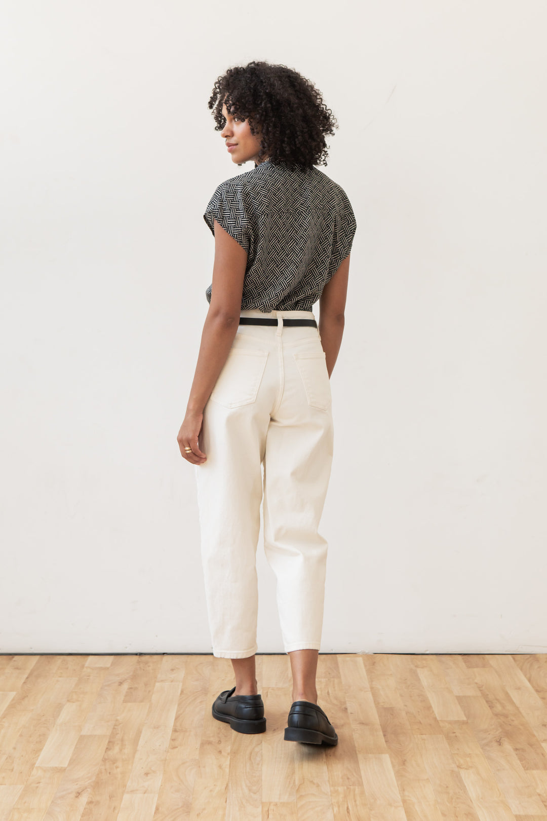 A person with curly hair stands indoors on a wooden floor, facing away. They wear the Bridge & Burn Ladd Shirt in Black Geo, light high-waisted pants with a black belt, and black shoes against a plain white wall.