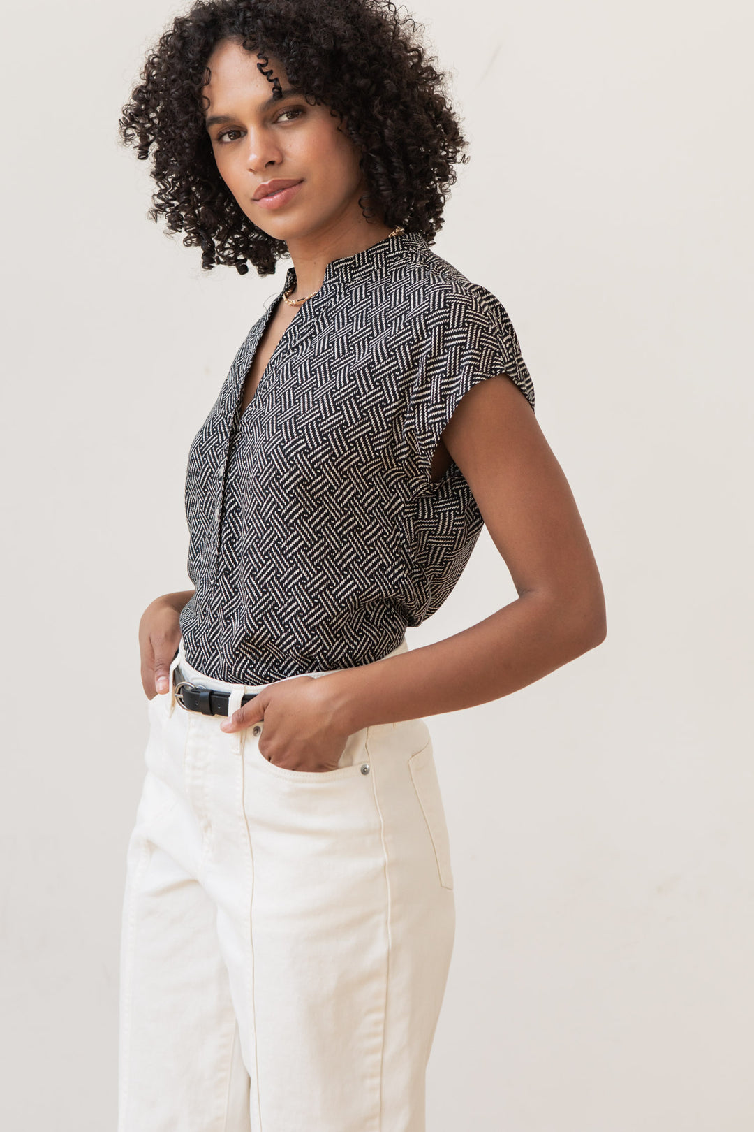 Woman with curly hair wears the Bridge & Burn Ladd Shirt in Black Geo, tucked into high-waisted white pants with a black belt, standing against a light background with one hand in her pocket.