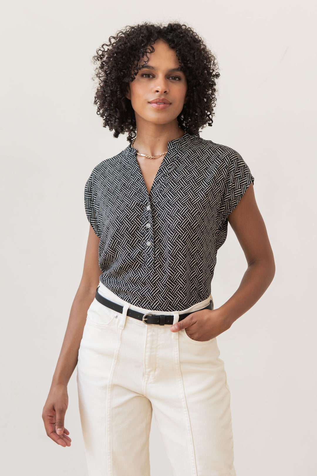 A woman with curly hair wears the Bridge & Burn Ladd Shirt in Black Geo, tucked into high-waisted off-white pants with a black belt, standing confidently against a plain light background.