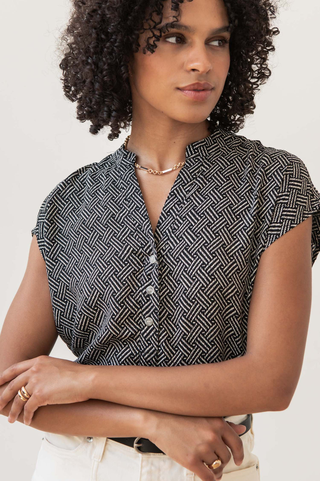 A woman with curly hair wears the Bridge & Burn Ladd Shirt in Black Geo, a gold necklace, and light pants. She stands with arms crossed, looking to the side against a plain background.
