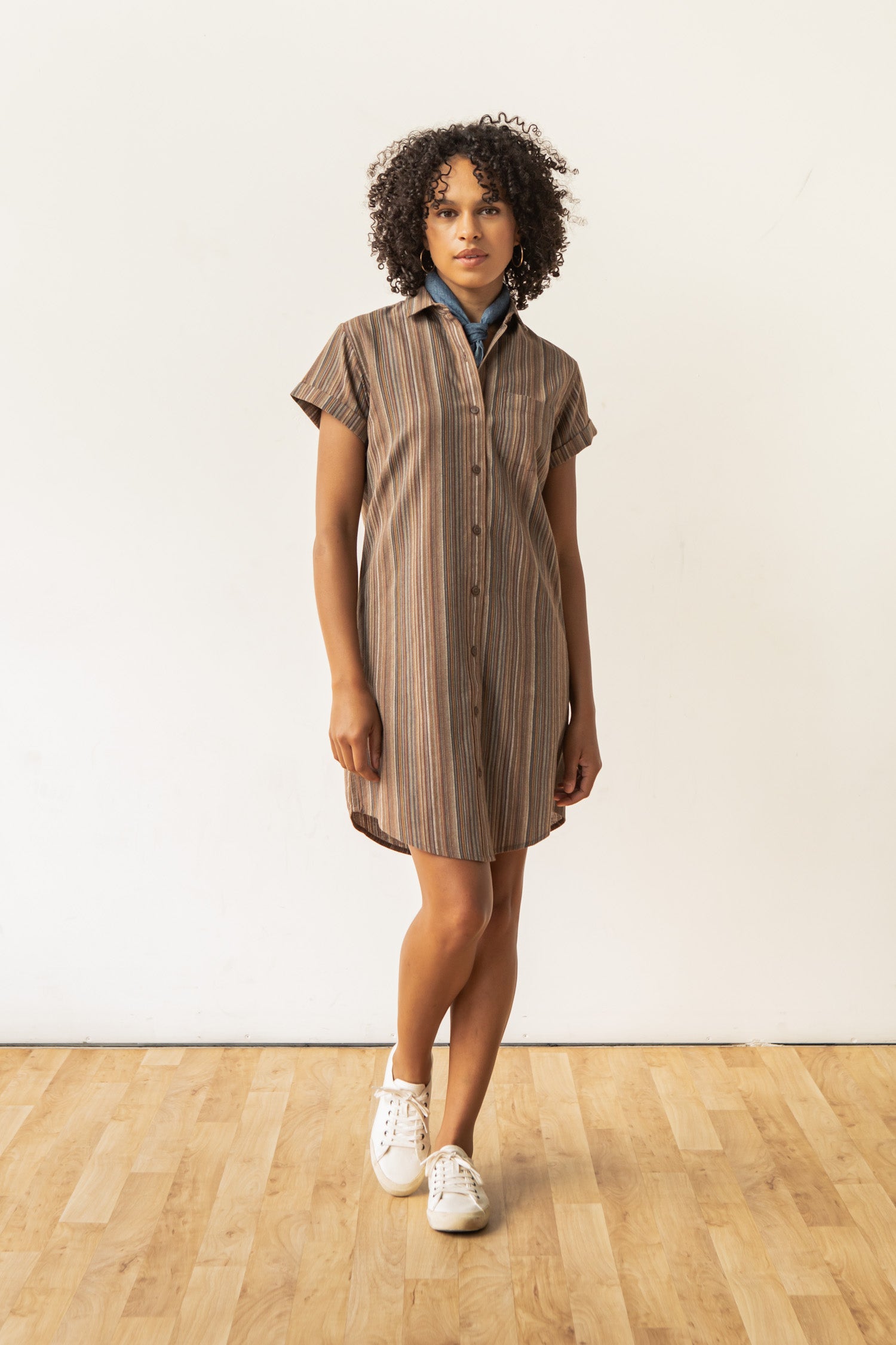 A person with curly hair wears the Bridge & Burn Loren Shirt Dress in Multistripe, paired with white sneakers and a blue scarf. They stand on a light wooden floor against a plain white wall.