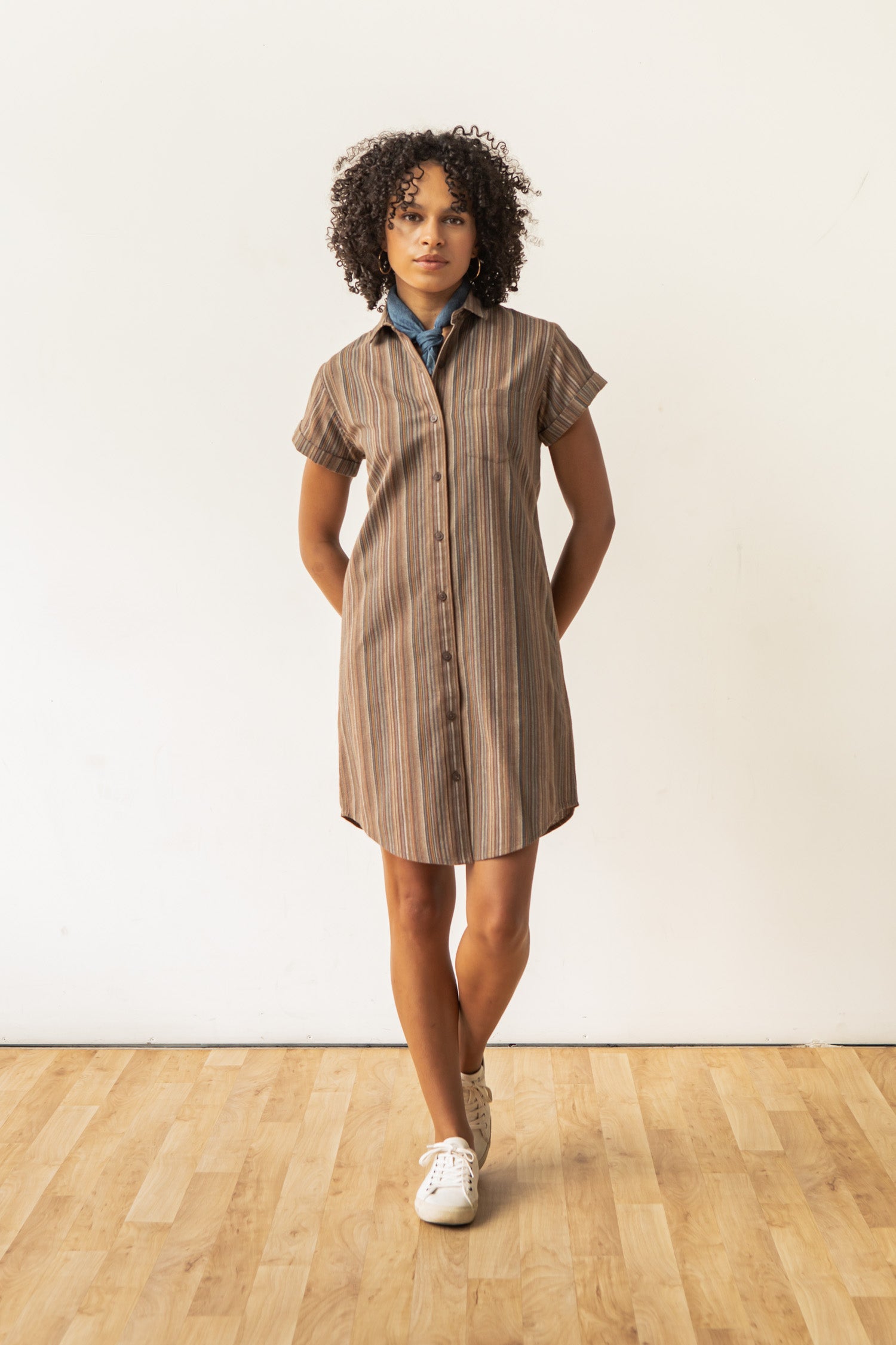 The Bridge & Burn Loren Shirt Dress / Multistripe is a short-sleeved, knee-length 100% cotton dress with brown, beige, and blue stripes, a collar, front buttons, and chest pocket from the Sustainable Series Collection. Laid flat on white.