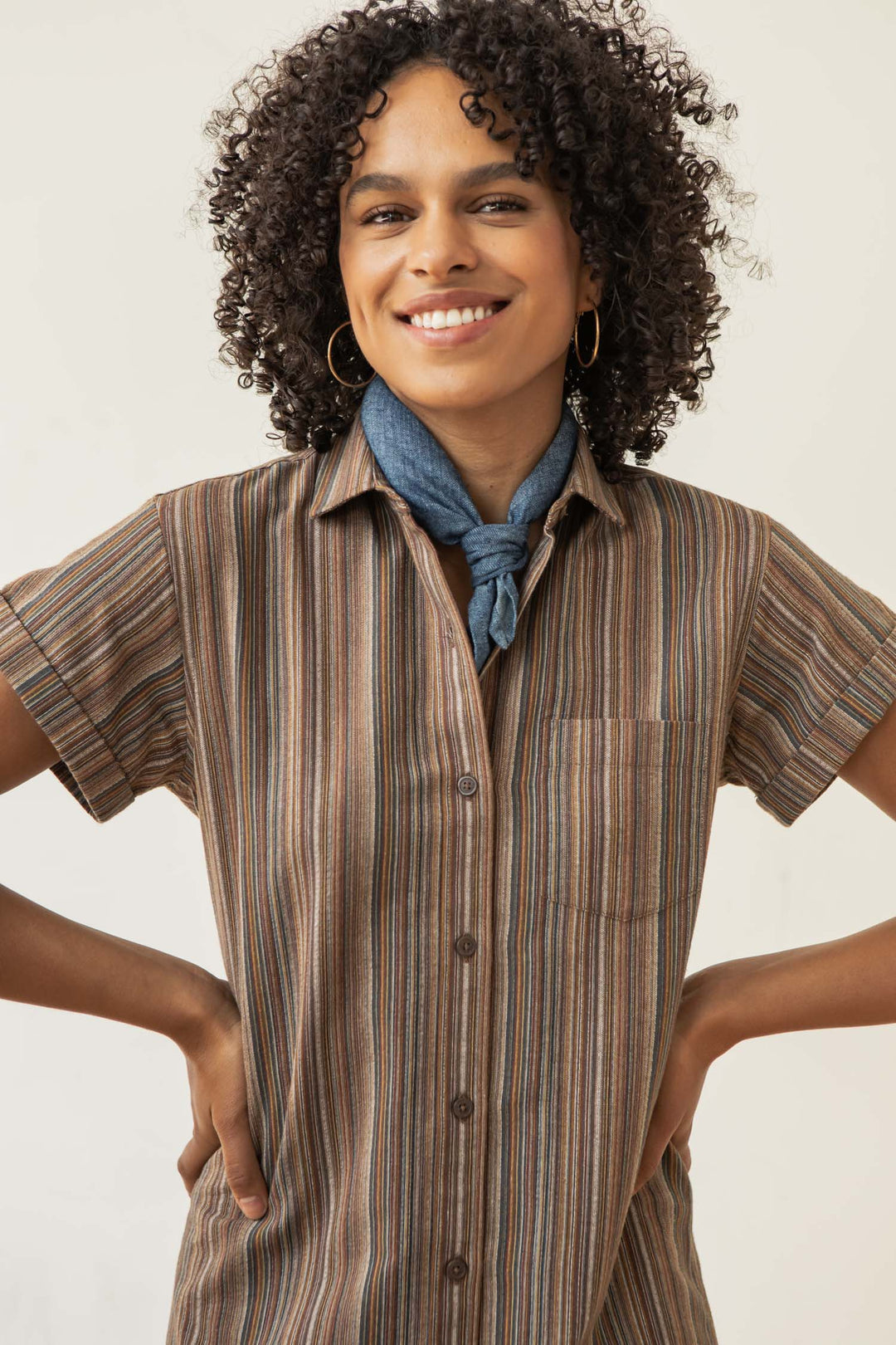 A smiling woman with curly hair wears the Bridge & Burn Loren Shirt Dress in Multistripe and a blue scarf, accessorized with gold hoop earrings. She stands hands on hips against a plain background.