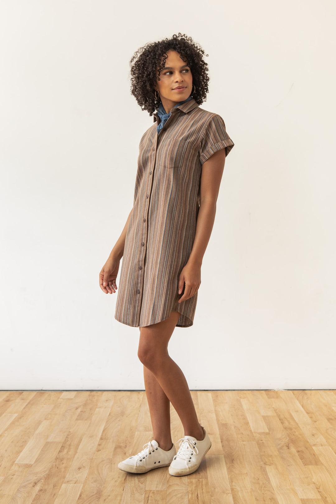 A person stands on a wooden floor wearing the Bridge & Burn Loren Shirt Dress / Multistripe with white sneakers. Their curly hair frames their face as they look slightly to the side in front of a plain white wall.