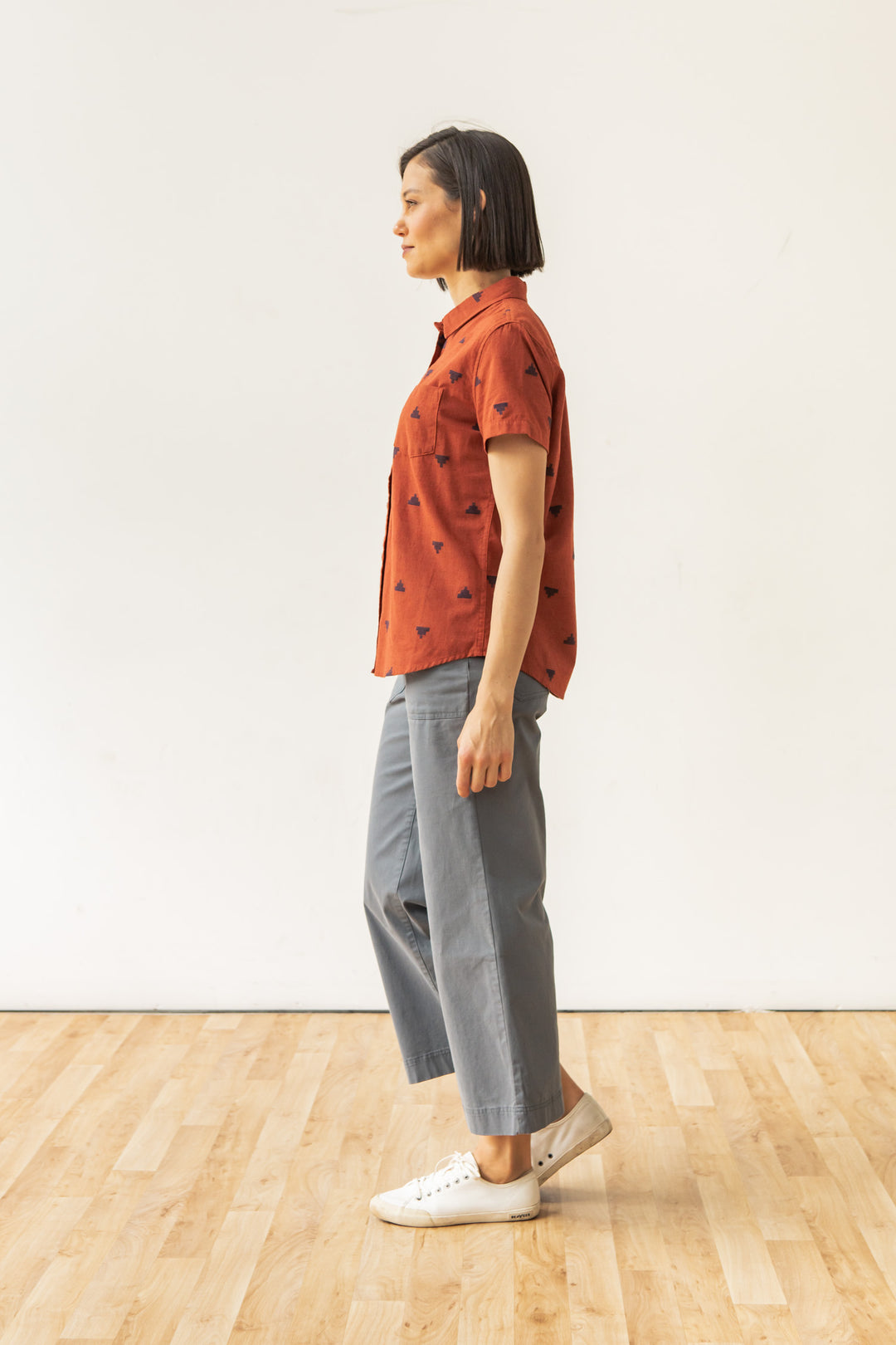 A person with short dark hair stands in profile on a wooden floor, wearing the Bridge & Burn Lana Shirt in Rust Fil-Coupé Pyramids, gray wide-leg pants, and white sneakers against a plain white background.
