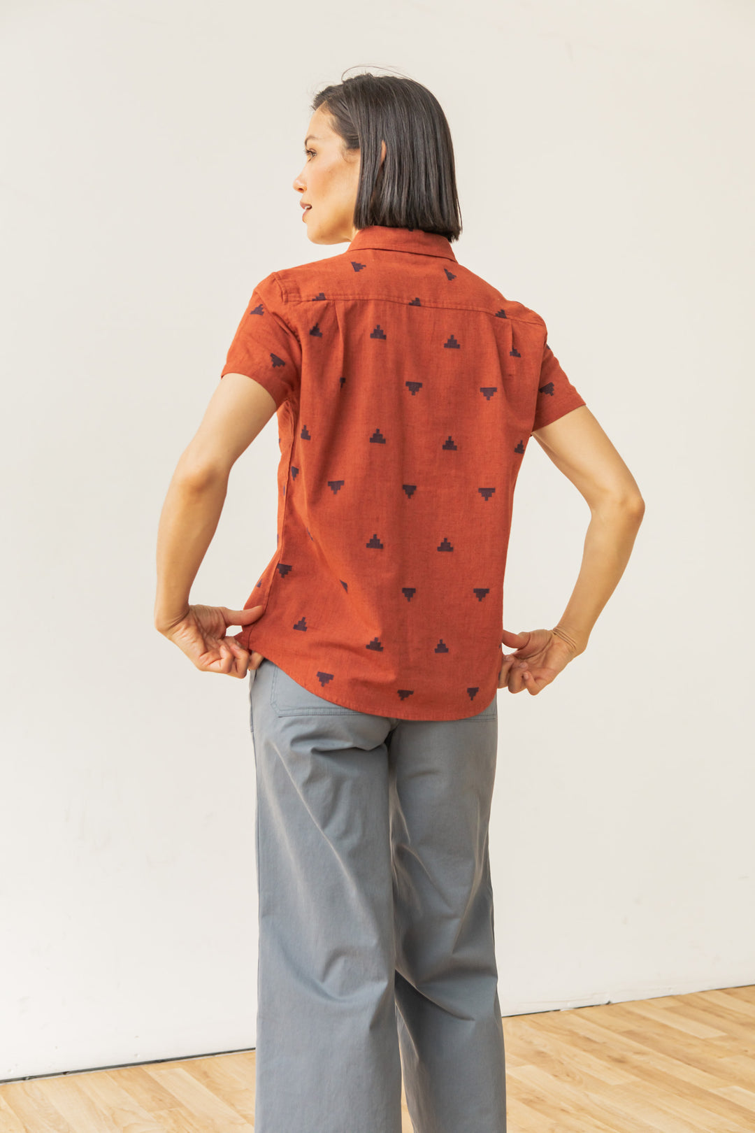 A person with short dark hair wears the Bridge & Burn Lana Shirt in Rust Fil-Coupé Pyramids and loose gray pants, standing indoors on a wooden floor facing away from the camera.