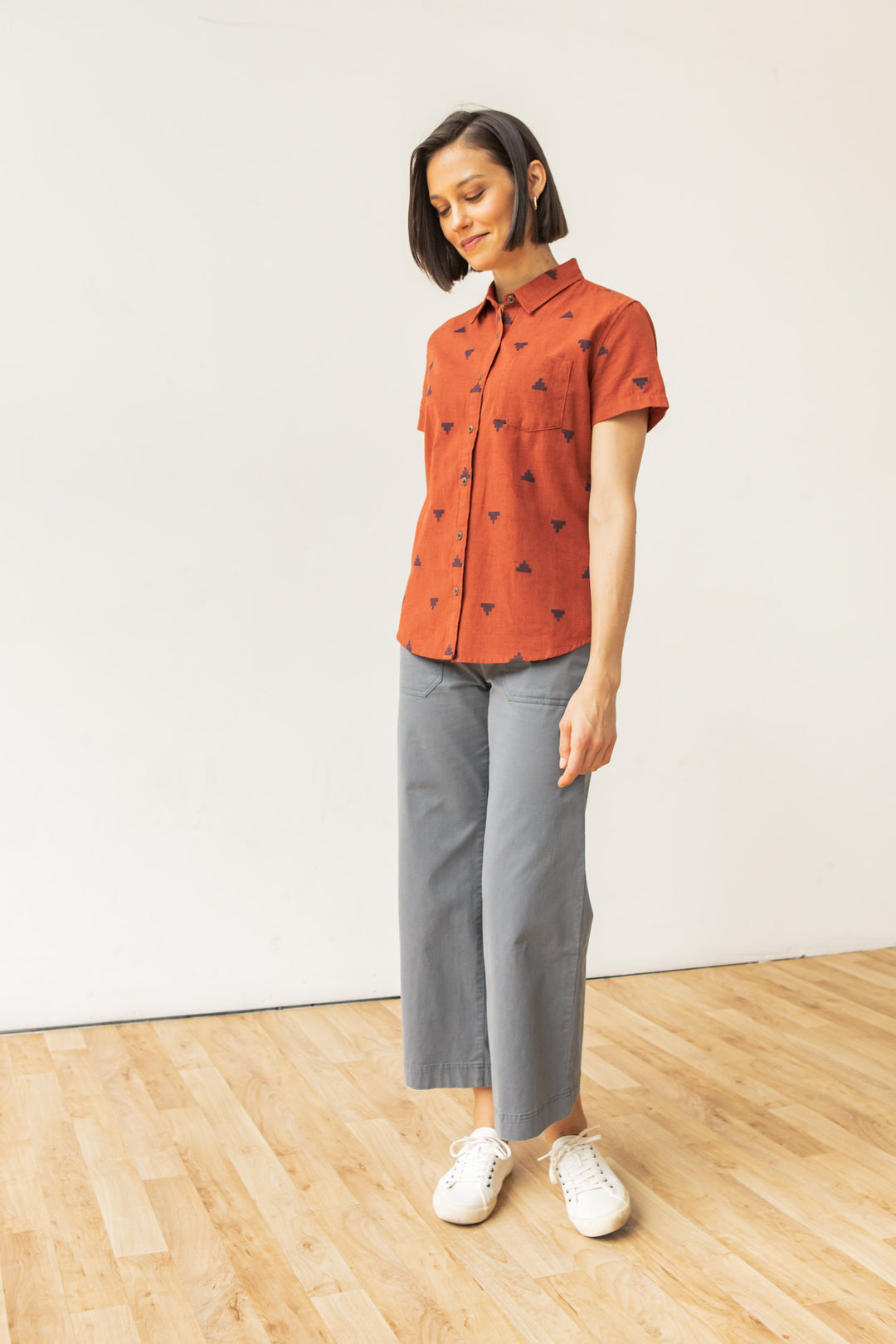 A woman with short dark hair stands on a wooden floor against a white wall. She wears the Bridge & Burn Lana Shirt in Rust Fil-Coupé Pyramids, gray wide-leg pants, and white sneakers, looking down with a slight smile.