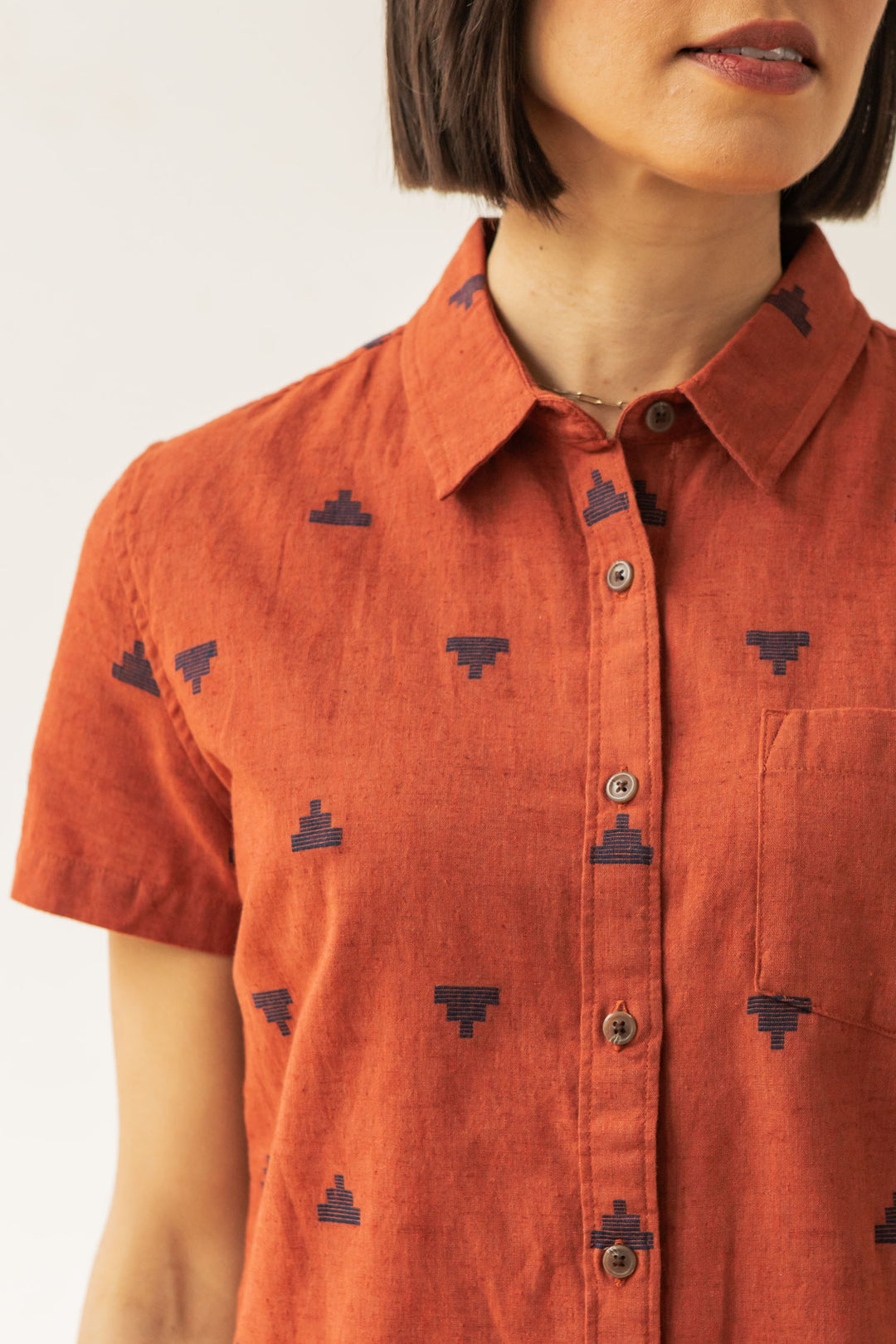 A woman wears the Bridge & Burn Lana Shirt in Rust Fil-Coupé Pyramids, a sustainable short sleeve style featuring a subtle navy geometric pattern. The photo shows her from shoulders to just above the waist; her hair is straight and chin-length.