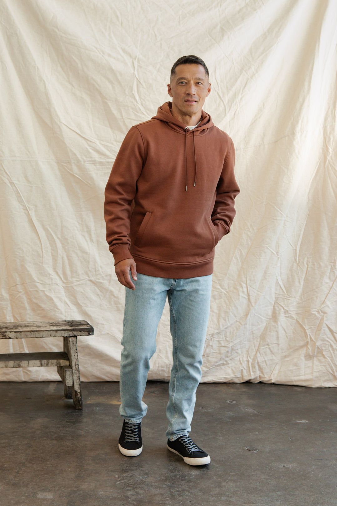 A man stands indoors on a concrete floor, wearing the Bridge & Burn Williams Pullover Hoodie in Burnt Umber with light blue jeans and black sneakers. One hand in his pocket, he poses by a rustic wooden bench before a beige fabric backdrop.