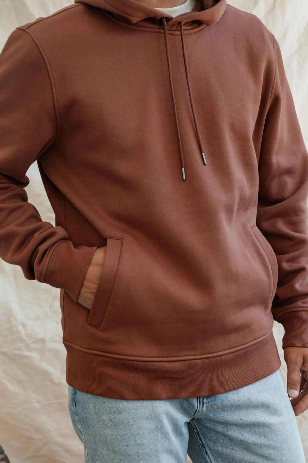 A person wearing the Bridge & Burn Williams Pullover Hoodie in Burnt Umber with a front pocket, paired with light blue jeans, stands against a light background. Their left hand is in the pocket and their face is not visible.