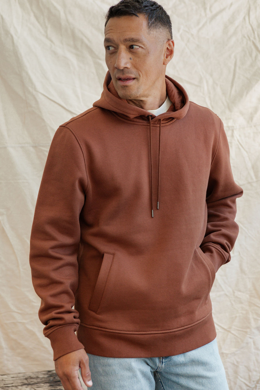 A person wearing the Bridge & Burn Williams Pullover Hoodie in Burnt Umber with the hood down and light blue jeans stands before a beige fabric backdrop, looking left with one hand in their pocket.