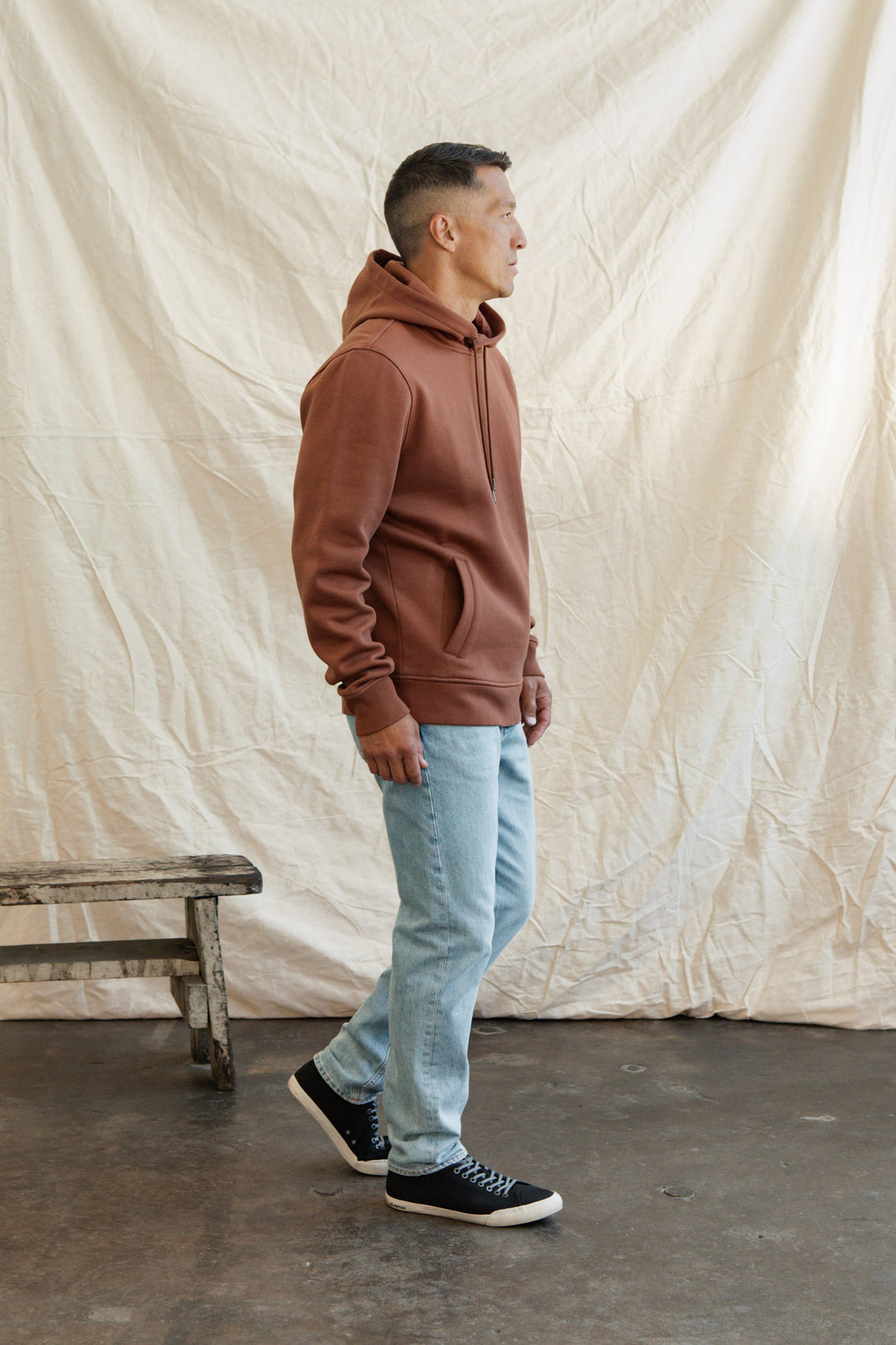 A man in the Williams Pullover Hoodie in Burnt Umber by Bridge & Burn, light blue jeans, and black sneakers stands indoors on a concrete floor beside a wooden bench, with a cream-colored fabric backdrop behind him.