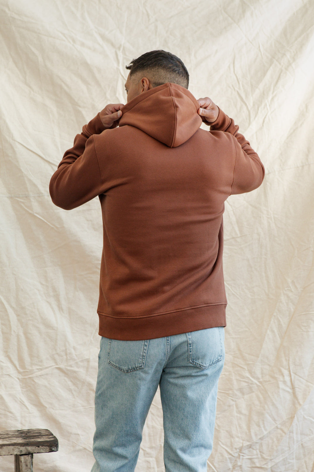 Wearing the Bridge & Burn Williams Pullover Hoodie in Burnt Umber and light blue jeans, a person stands with their back to the camera, adjusting the hood in front of a beige fabric backdrop.