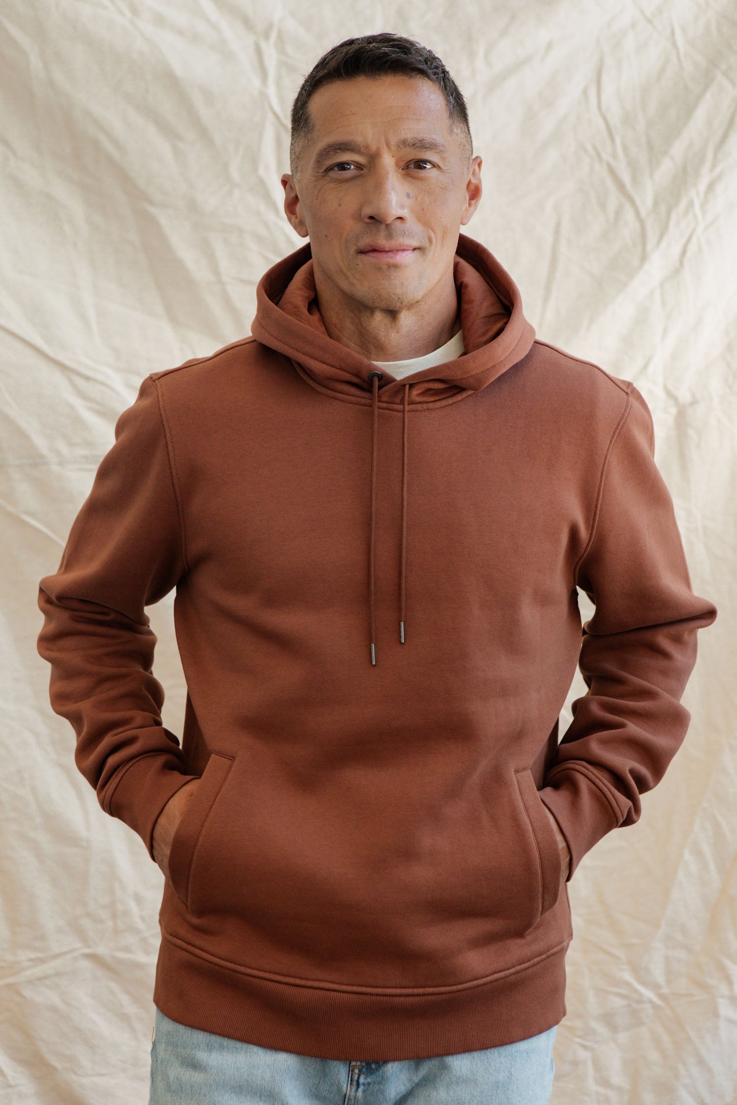 A man with short hair and a slight smile faces the camera, hands in the front pockets of the Bridge & Burn Williams Pullover Hoodie in Burnt Umber. The background is a light, textured fabric.
