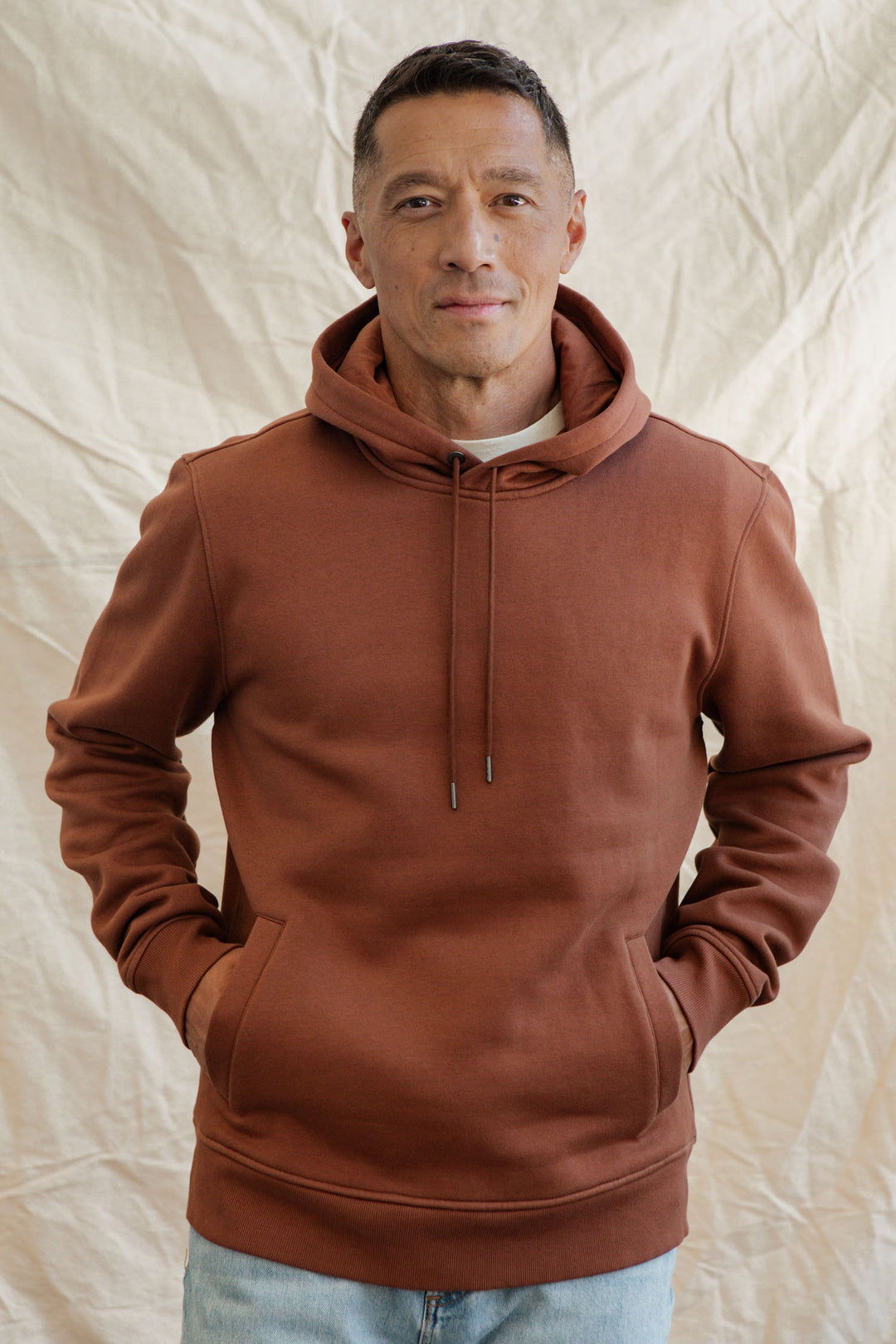A man with short hair and a slight smile faces the camera, hands in the front pockets of the Bridge & Burn Williams Pullover Hoodie in Burnt Umber. The background is a light, textured fabric.