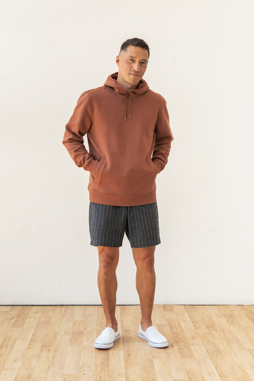 A man stands indoors on a wooden floor, wearing the Bridge & Burn Williams Pullover Hoodie in Burnt Umber. With short dark hair and hands in his hoodie pockets, he faces the camera against a plain white wall.