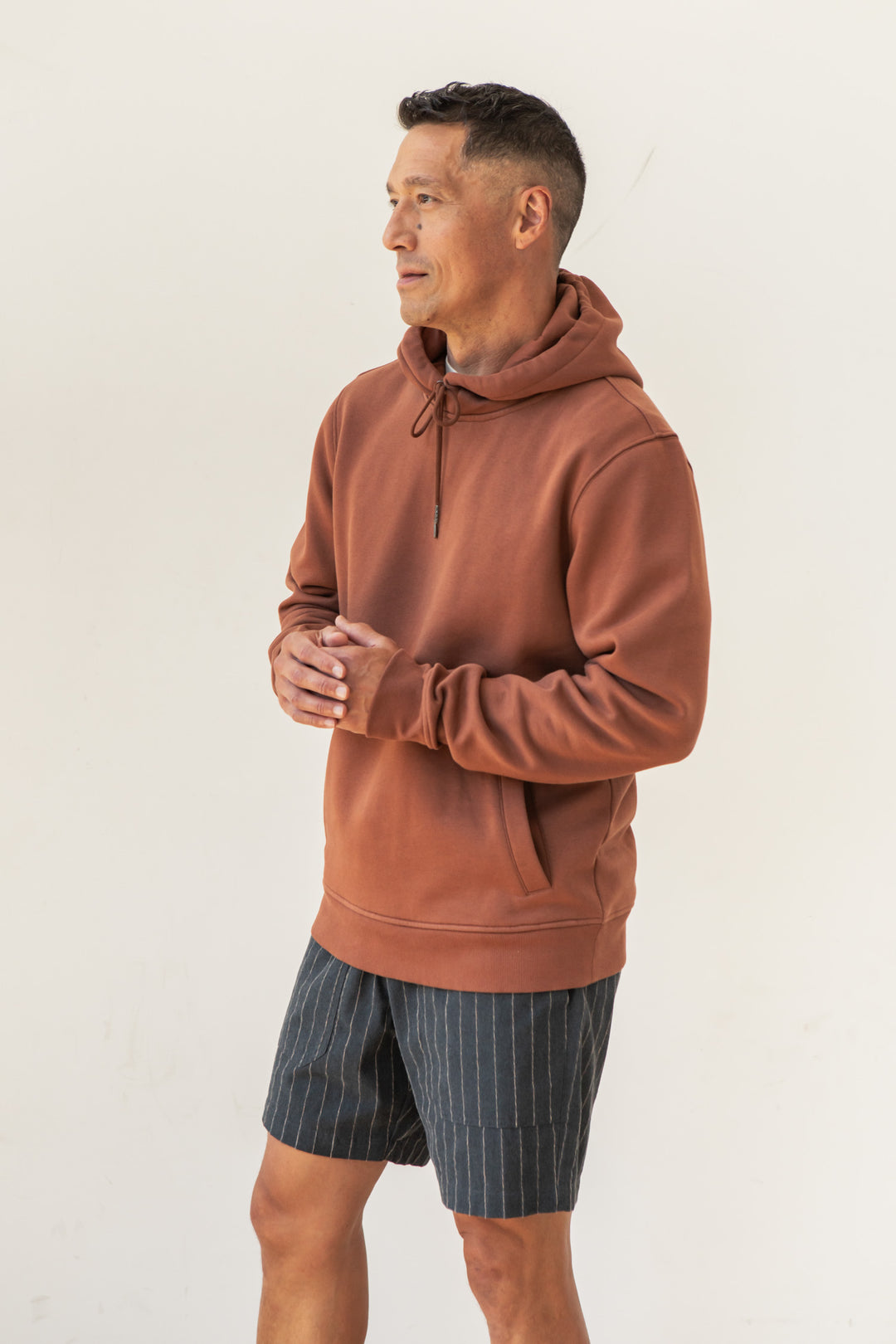 A man wearing the Bridge & Burn Williams Pullover Hoodie in Burnt Umber and pinstriped shorts stands against a plain white background, looking slightly to the side with his hands clasped together.