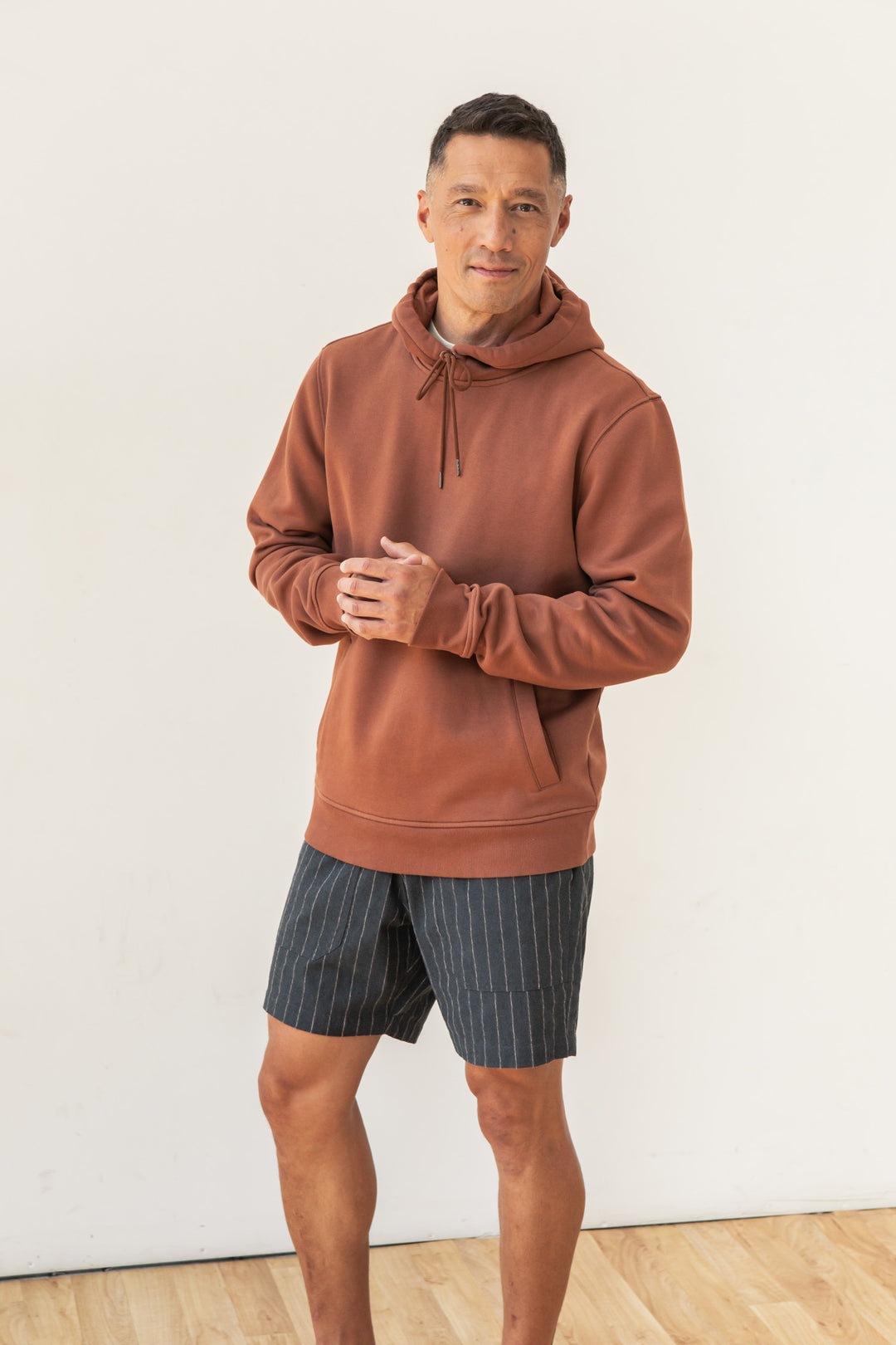 A man stands indoors against a white wall, looking at the camera and gently clasping his hands. He wears the Bridge & Burn Williams Pullover Hoodie in Burnt Umber with gray pinstriped shorts. The light wood floor completes the scene.