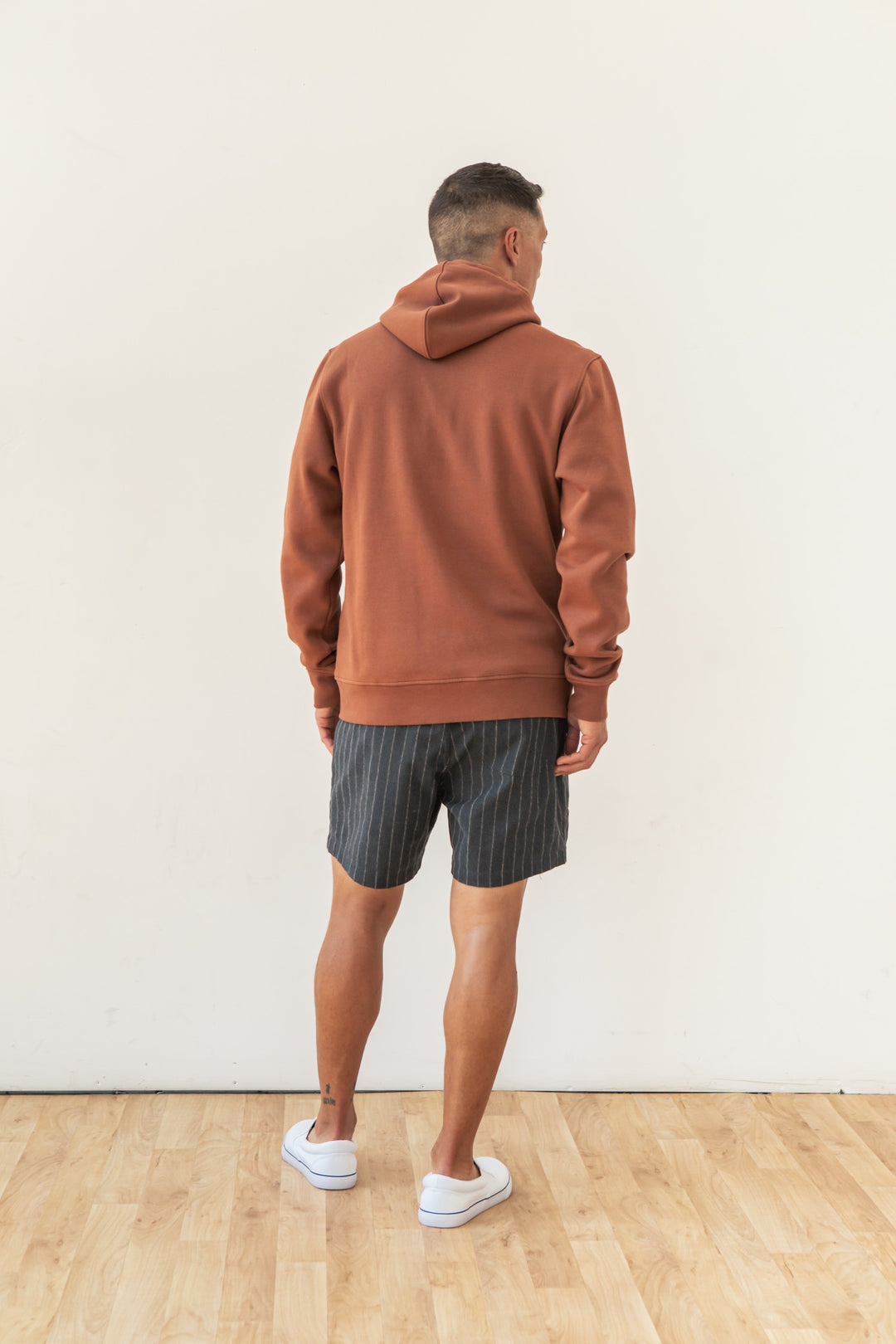 A man stands indoors on a wooden floor facing away from the camera, wearing the Bridge & Burn Williams Pullover Hoodie in Burnt Umber with gray pinstriped shorts and white slip-on shoes against a plain white wall.