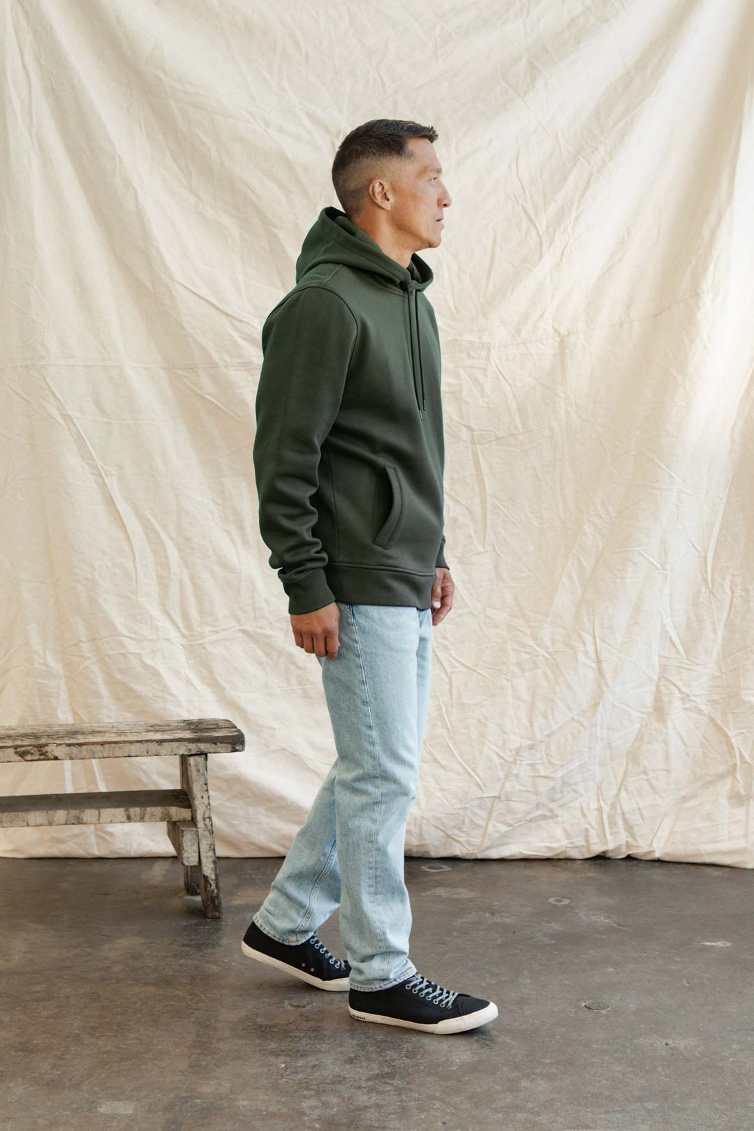 A man in the Bridge & Burn Williams Pullover Hoodie in Forest Night, light blue jeans, and black sneakers stands sideways on a concrete floor before a crumpled beige fabric backdrop, with a rustic wooden bench to his left.