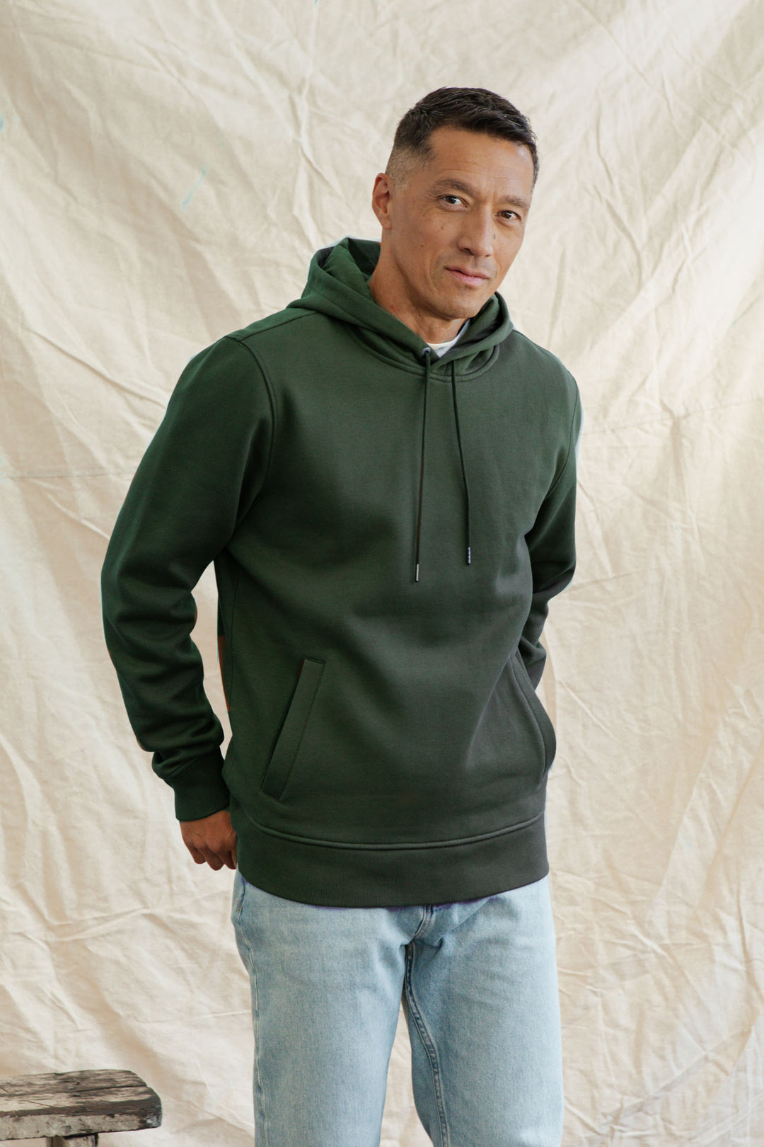 A man wearing the Bridge & Burn Williams Pullover Hoodie in Forest Night and light blue jeans stands before a beige, textured backdrop, looking at the camera with a slight smile.