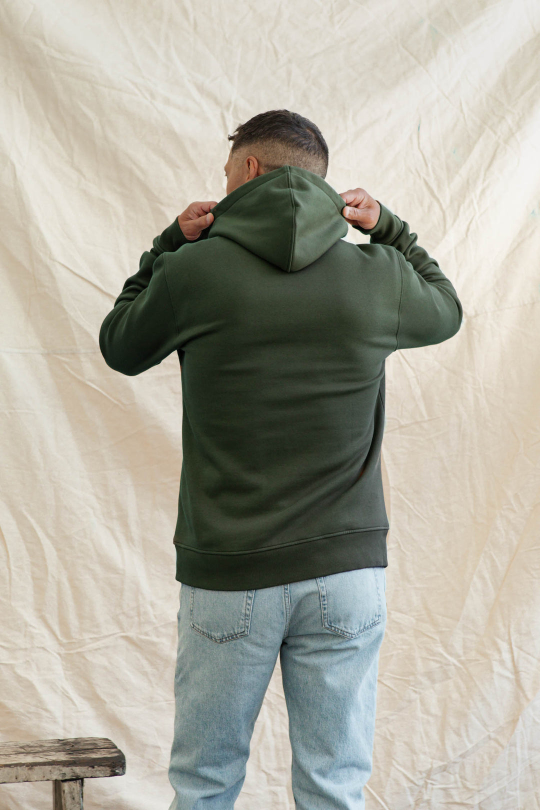 A person wearing the Bridge & Burn Williams Pullover Hoodie in Forest Night and light blue jeans stands facing away, holding the hood with both hands against a beige, slightly wrinkled fabric backdrop.