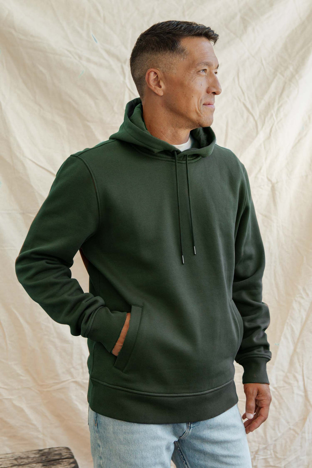 A man wearing the Bridge & Burn Williams Pullover Hoodie in Forest Night and light blue jeans stands against a beige, wrinkled fabric backdrop, one hand in his pocket as he looks to the side.
