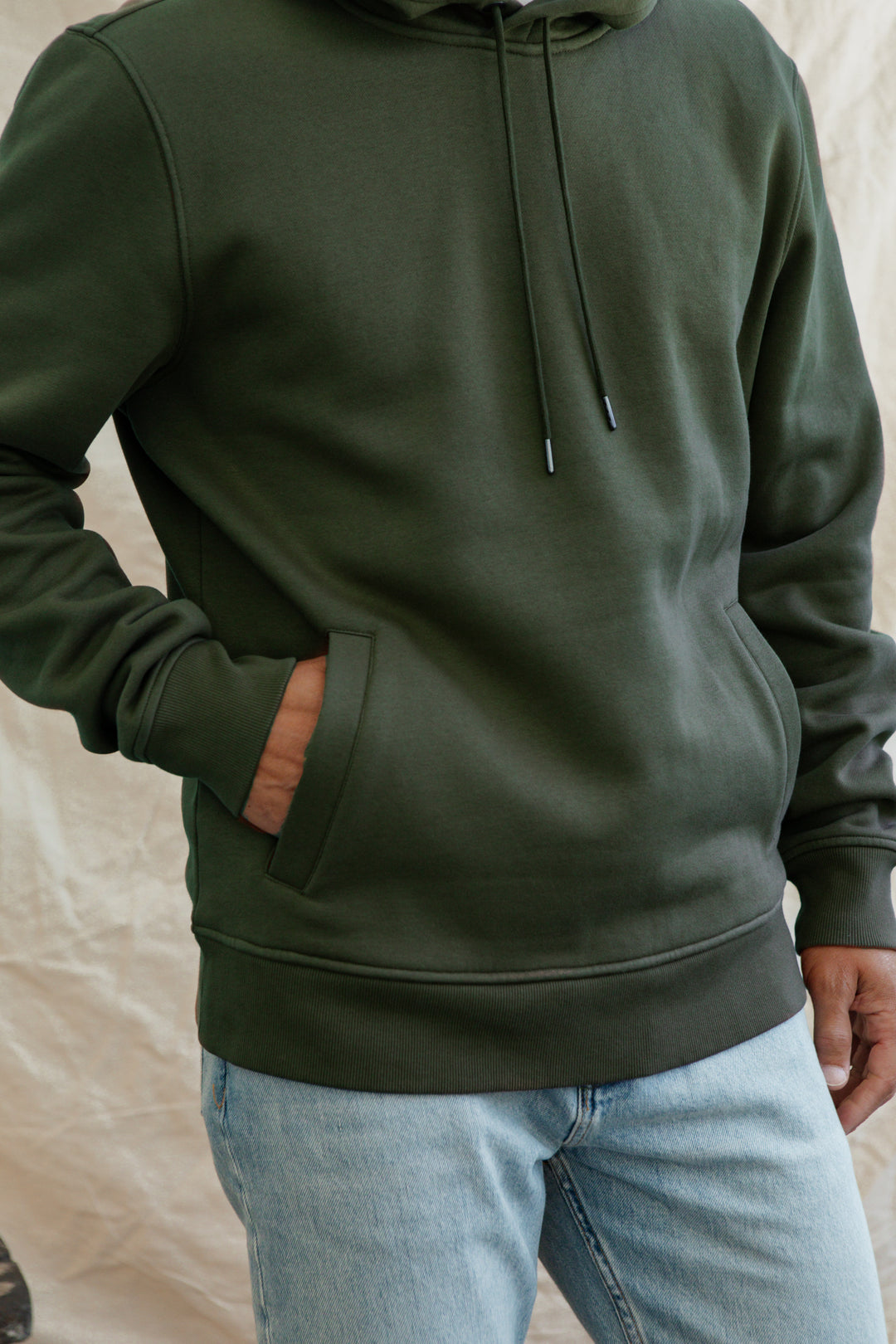 A person wears the Bridge & Burn Williams Pullover Hoodie in Forest Night (dark green recycled polyester) with one hand in the pocket and light blue jeans, standing against a light textured background. Their head is not visible.