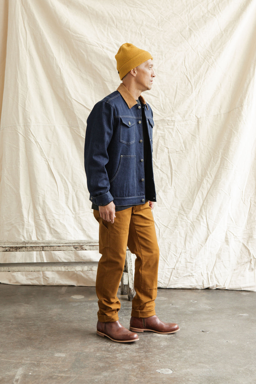 Indoors on concrete, a person wears a yellow beanie, denim jacket, and Bridge & Burn’s Wallace Utility Pant in Hickory Canvas with tool pockets. Brown boots finish the look as they gaze right before a cream fabric backdrop.
