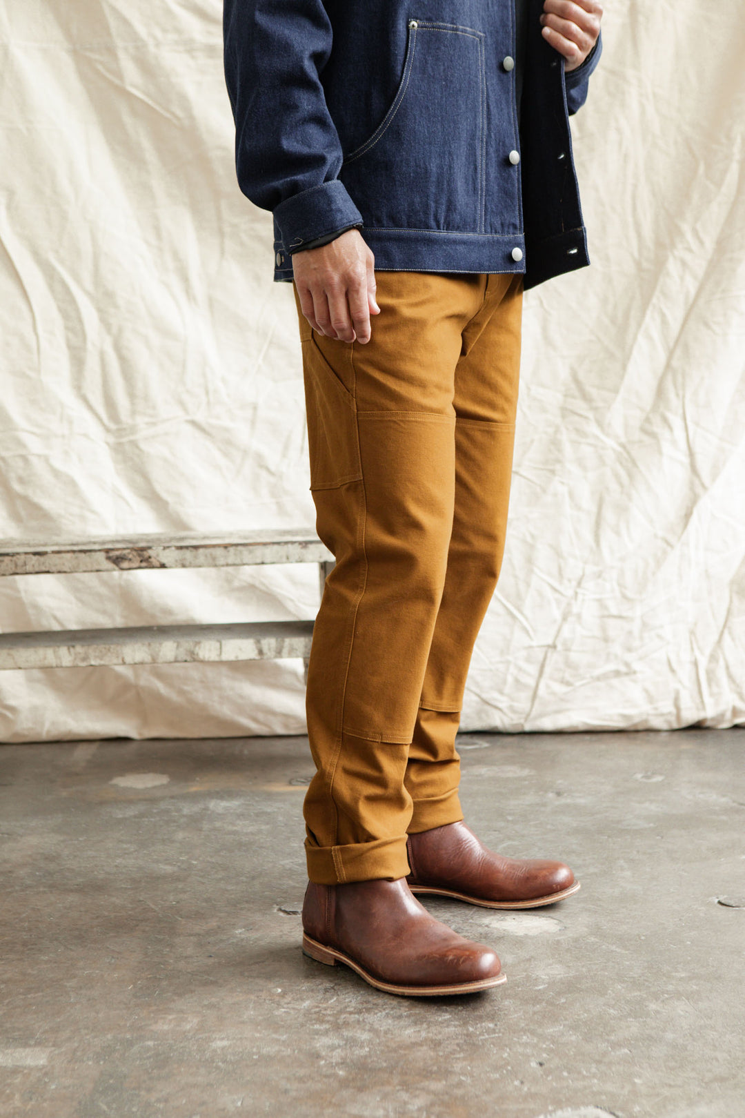 Indoors, a person wears Bridge & Burn’s Wallace Utility Pant in Hickory Canvas with a dark denim jacket and brown boots. The background has a beige fabric drape and wooden bench; the upper face is not visible.