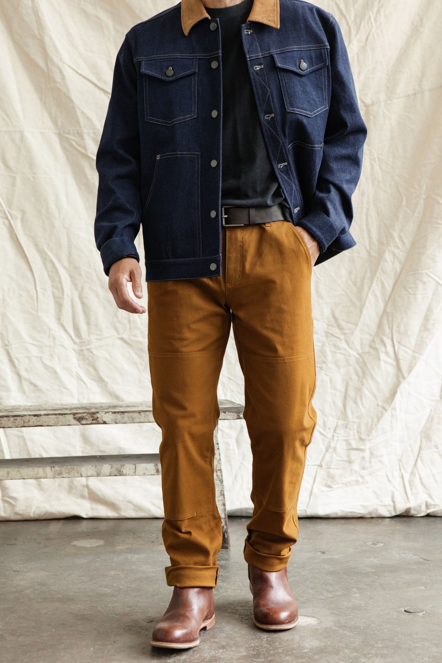 A person in a dark denim jacket, black t-shirt, Bridge & Burn Wallace Utility Pant in Hickory Canvas, and brown leather boots stands indoors before a cream fabric backdrop. The person’s face is not visible.